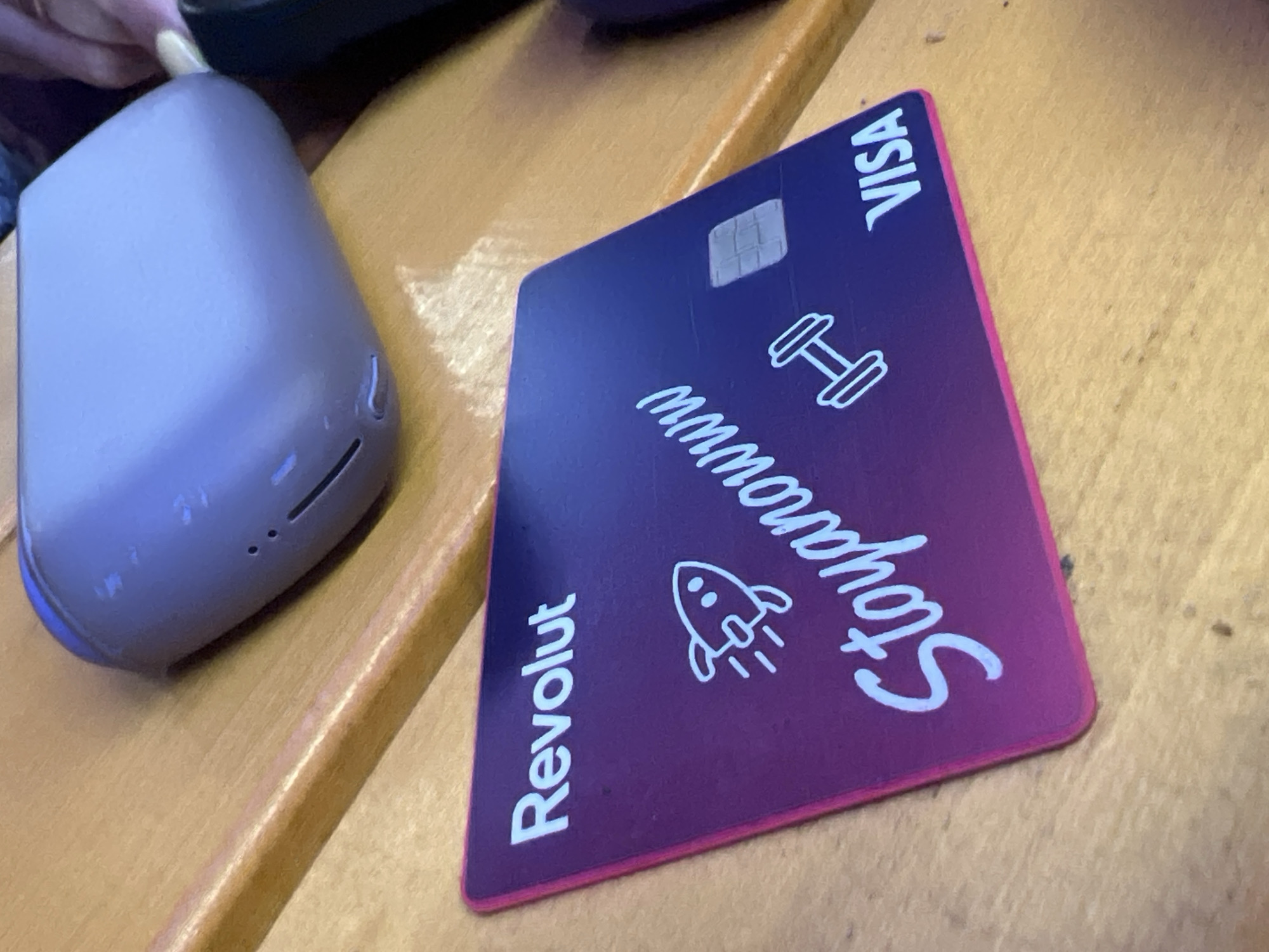 The image captures a close-up view of a purple and pink gradient Revolut Visa card alongside a grey electronic device, both resting on a light brown wooden surface.
The credit card is the central focus, exhibiting a gradient design that fades from dark purple at the top to a vibrant pink towards the bottom. The brand name "Revolut" is printed in white text along the bottom-left edge, while "VISA" is visible vertically in white text at the top-right corner. A standard silver card chip is embedded in the top-left section. Decorative white outline icons include a rocket ship on the left side and a dumbbell on the right. Across the center, the name "Stoyanowww" is written in a distinct white, handwritten-style font. The card itself has a bright pink edge.
To the left of the card is a smooth, rounded rectangular electronic device, possibly a portable charger or an earbud case, in a muted grey or lavender color. This device shows noticeable wear and tear, with several scratches and scuff marks visible on its surface. It features a small, elongated opening and two tiny circular indicator lights on one side. A manicured fingernail, with light polish, is partially visible at the very top-left of the frame, near the device.
The items are situated on a wooden surface that has a visible grain and a light, warm brown tone, suggesting an indoor setting like a desk or table. No specific activity is occurring; the objects appear to be casually placed.