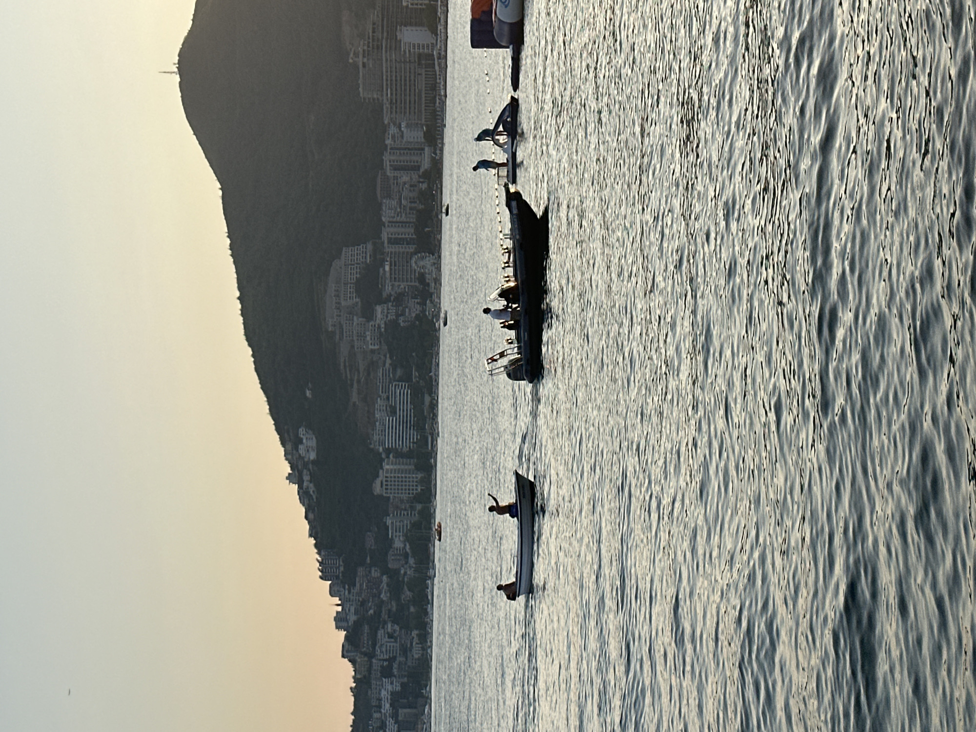 The image depicts a serene coastal scene at sunset in Budva, Montenegro. In the foreground, the calm waters of the Adriatic Sea reflect the soft, warm hues of the twilight sky. Several boats are visible on the water. A small, white rowboat with two silhouetted figures navigates the sea, one appearing to be rowing or steering. Slightly further to the right, a larger, darker boat with multiple people on board is also present. In the background, the city of Budva sprawls across the hillside, characterized by a dense collection of buildings, some appearing to be hotels or apartments, with a prominent hill forming the backdrop. The peak of the hill is marked by a telecommunications tower. The overall atmosphere is peaceful and picturesque, capturing a moment of leisure and natural beauty.