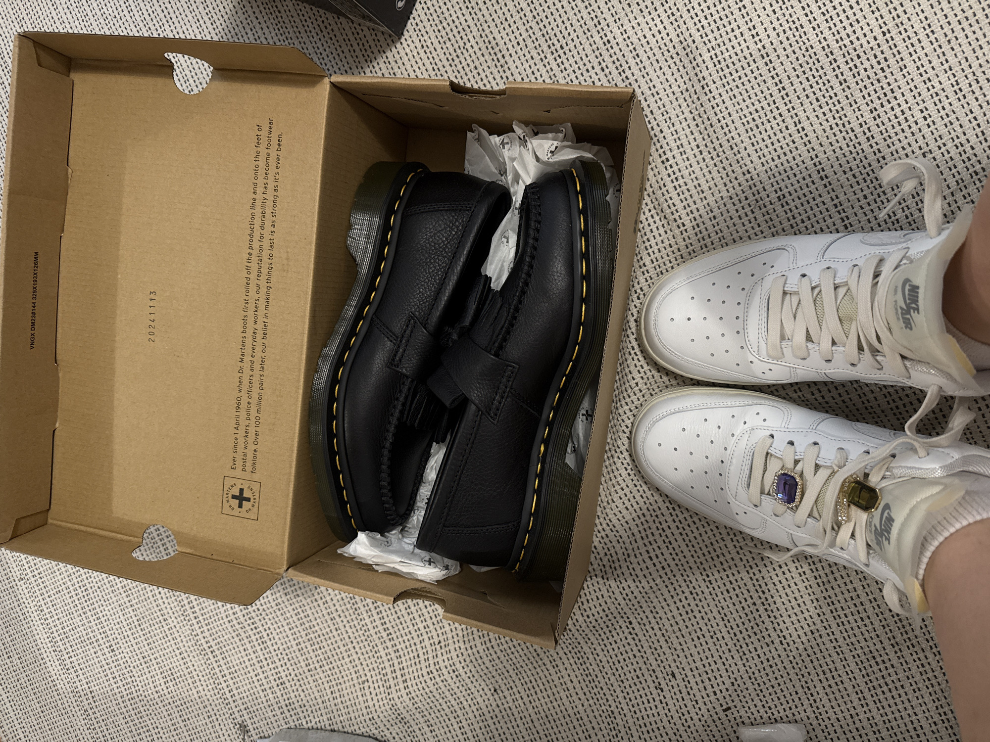 The image presents an indoor scene, likely in a home in Hainburg an der Donau, Austria, showcasing a pair of new black Dr. Martens loafers inside their cardboard box, positioned on a light-colored, textured carpet. Alongside the box, a person's lower legs and feet are visible, wearing white Nike Air Force 1 sneakers.
The open Dr. Martens shoebox, made of brown cardboard, contains the black loafers. They feature a textured leather finish, traditional yellow stitching along the sole, and a tassel detail on the vamp. Inside the box, the shoes are partially wrapped in white tissue paper. Printed text on the box includes "VNGX DM23#144 329X193X126MM" and "20241113". A longer text block reads: "Ever since 1 April 1960, when Dr. Martens boots first rolled off the production line and onto the feet of postal workers, police officers and everyday workers, our reputation for durability has become footwear folklore. Over 100 million pairs later, our belief in making things to last is as strong as it's ever been." The Dr. Martens logo is also visible.
The white Nike Air Force 1 sneakers worn on the feet display the "NIKE AIR" branding on the side and feature small perforations on the toe box. Each sneaker is accessorized with a prominent, rectangular, jewel-like lace charm; one is a deep blue and the other a yellow/gold, set with smaller clear stones. The overall activity depicted suggests the unboxing or presentation of the new Dr. Martens loafers, contrasted with the worn but stylized Nike sneakers. The lighting indicates an indoor setting, with no specific time of day or weather discernible.