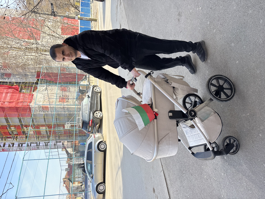 A man, appearing to be in his 30s or 40s with short dark hair, stands on a paved surface, looking directly at the camera. He is dressed in a black quilted hooded jacket, a white t-shirt, black athletic pants, and black athletic shoes. He holds the handle of a light beige or off-white baby stroller, which features a large bassinet, black wheels, and a light-colored frame. A small Bulgarian flag with white, green, and red horizontal stripes is attached to the right side of the stroller's bassinet. A matching light-colored bag hangs from the stroller's handle.
In the midground, behind the man and stroller, an urban street is visible with two parked vehicles: a silver or light gold Alfa Romeo 159 Sportwagon and a dark-colored Mercedes-Benz ML-Class SUV. Behind the vehicles and a chain-link fence, part of a sports court with a basketball hoop structure is discernible.
The background is dominated by a multi-story building under construction or renovation, heavily draped in scaffolding, red safety netting, and white protective sheeting, with "300" visible in repeating patterns on the sheeting. To the left of this structure, residential buildings with sloped, tiled roofs are visible. Bare trees and a blue barrier are present on the far right. The sky is clear and blue, suggesting a bright day in Byala Slatina, Bulgaria.