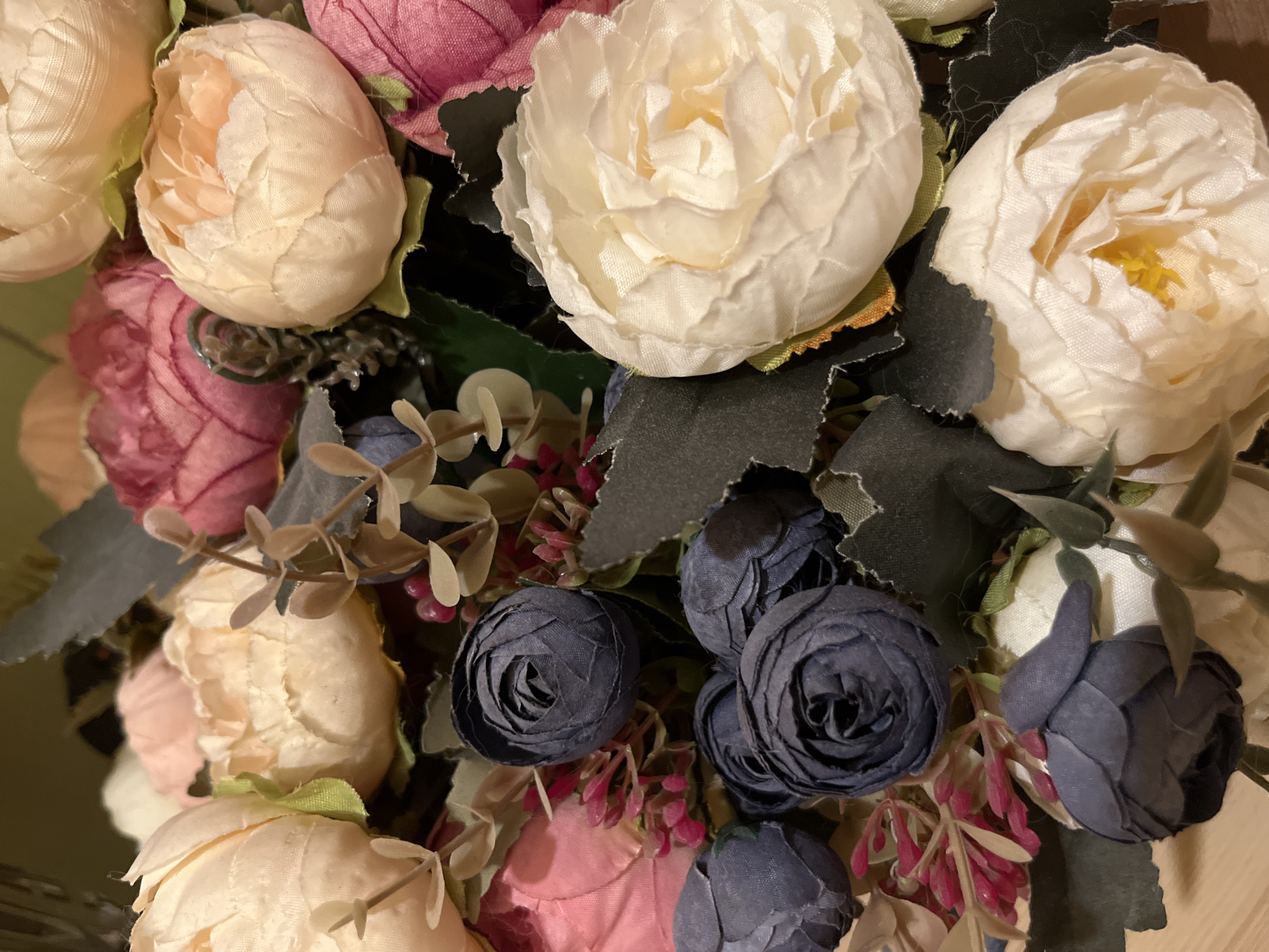 A close-up, overhead shot features a bouquet of artificial flowers, predominantly in shades of cream, peach, pink, and dark blue. The flowers have layered petals, creating a textured, romantic aesthetic. Accompanying the blooms are dark green artificial leaves and small, dark red berries on thin branches. The bouquet is presented in an unspecified location within the city of Ivanteyevka, Russia, with a wooden surface visible at the bottom of the frame, suggesting a tabletop or similar setting. There are no people or animals present in the image.