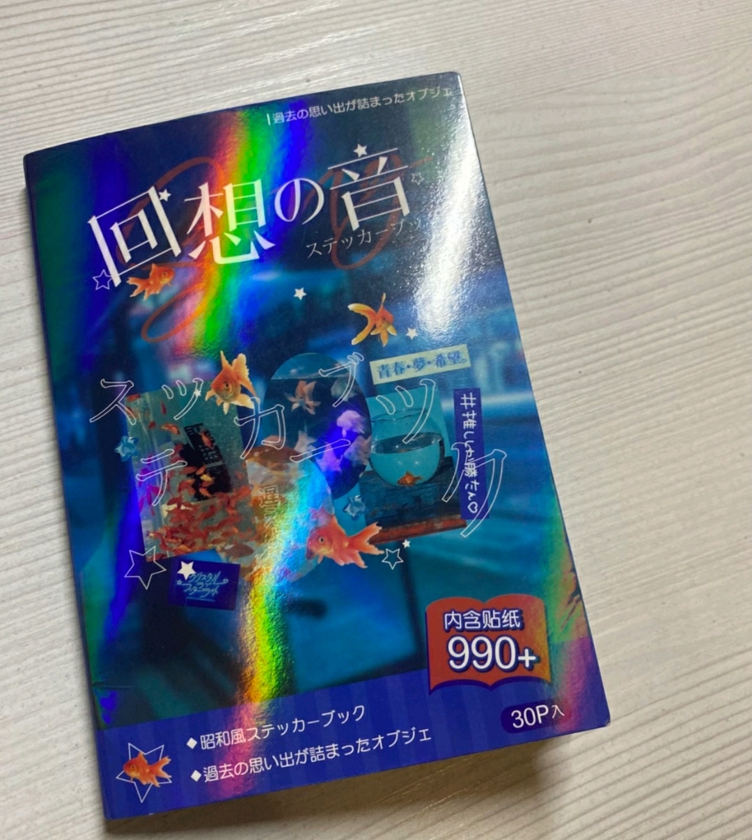 The image shows a sticker book titled "Echoes of Memories" with Japanese text. The cover features goldfish, bowls, and stars against a blue background with holographic effects. The text indicates it contains 990+ stickers and is reminiscent of the Showa era. The image is taken indoors on a light-colored surface.
