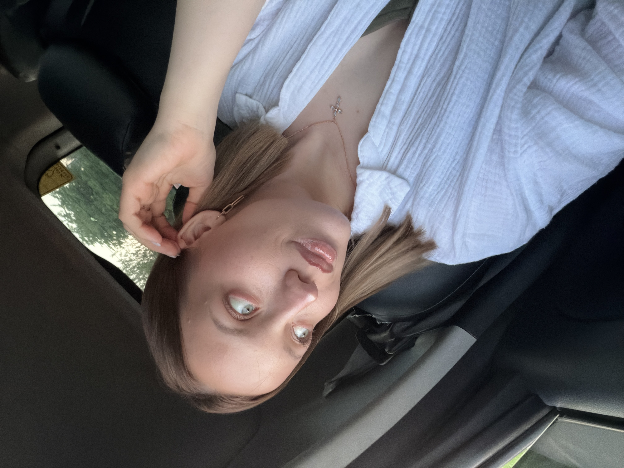 A woman with light brown hair and fair skin is seated in a car. She is wearing a white shirt and a delicate necklace with a cross pendant. She has one hand raised to her ear. The setting appears to be inside the car, with hints of greenery visible through the window, suggesting an outdoor location. The scene is an interior car setting with an individual posing within. There is no verifiable evidence within the image to confirm its location as Reshetnikovo, Russia.