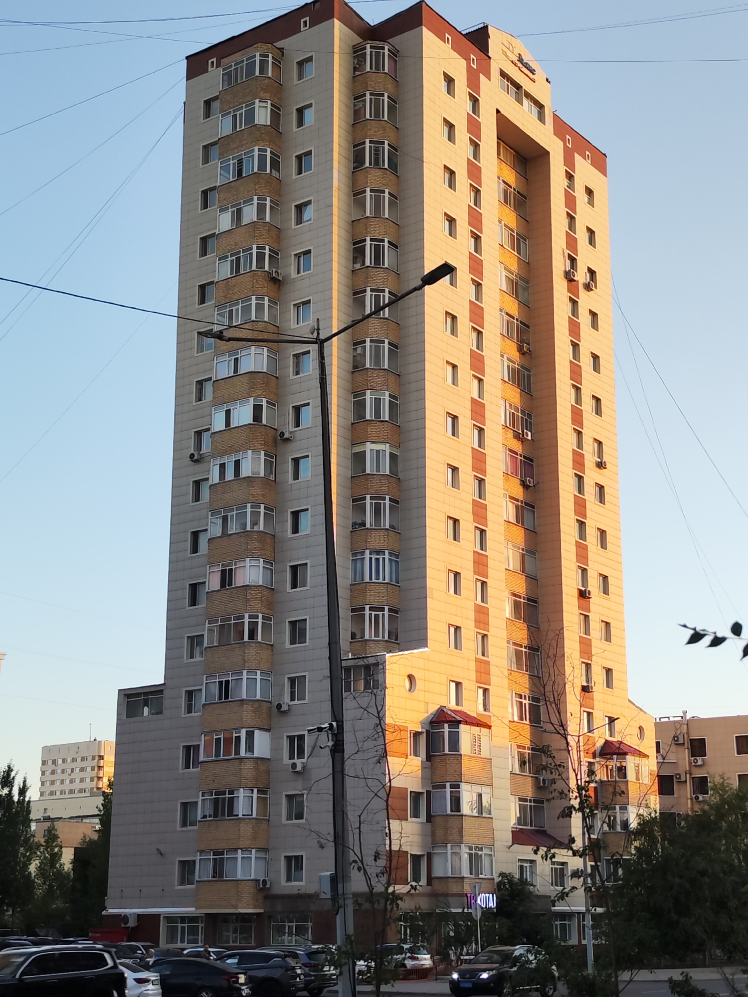 A tall, modern apartment building with a beige and red facade stands in a city, likely Astana, Kazakhstan, under a clear blue sky during daylight. The building features numerous windows and balconies, many of which are recessed and have a curved, bay-window design. The lower section of the building is clad in gray panels. In the foreground, several vehicles are parked, including black SUVs and sedans. Trees and power lines are visible around the building. There is a street lamp in front of the building. In the background, another, lighter-colored building with a similar window arrangement can be seen.
