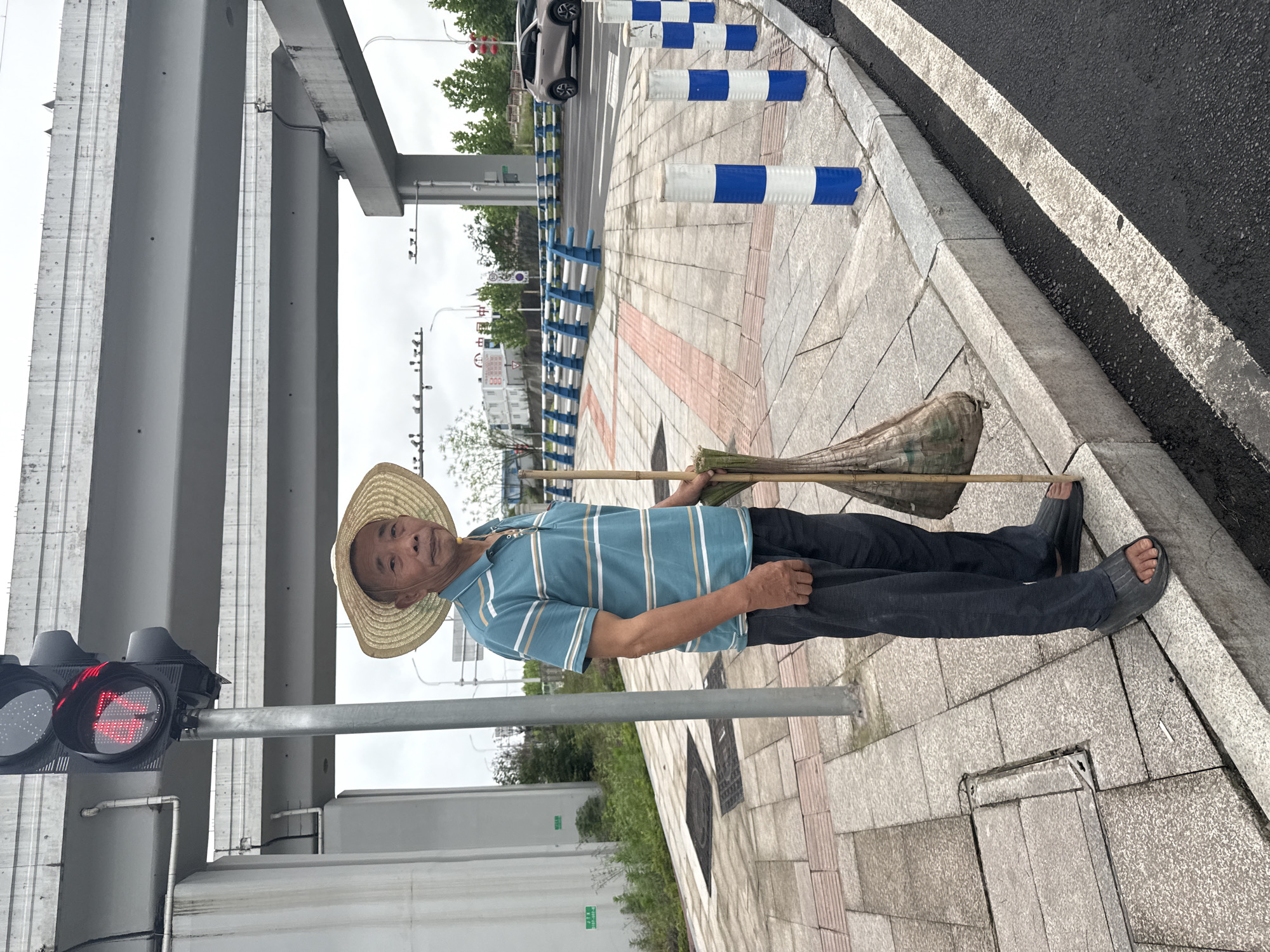 A man stands on a sidewalk in Jiangjin, China. He wears a straw hat, a blue and white striped shirt, dark pants, and sandals. He holds a bamboo broom. A traffic light next to him shows a red pedestrian signal. Above, there is an overpass. Blue and white traffic barriers line the road. The sky is overcast.