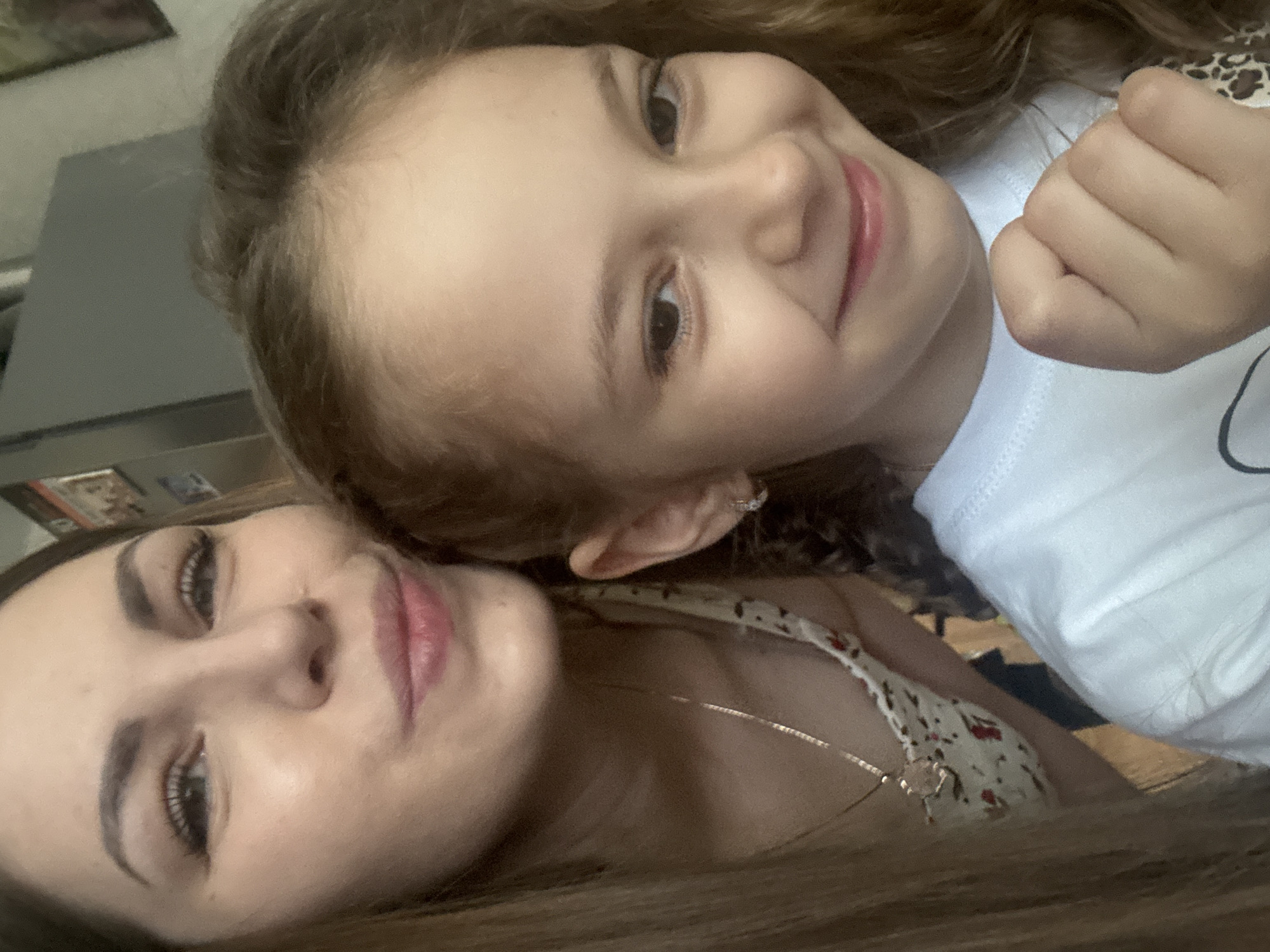 The image shows two people lying down, likely taking a selfie. The person in the foreground appears to be an adult female with brown hair, fair skin, and wearing a floral top and a necklace. The other person is a child with light brown hair, fair skin, wearing a white shirt, and is smiling. The background is blurry, but a grey object resembling a cabinet or appliance is visible. No information is available to confirm that the image was taken in Раздолье, Russia.
