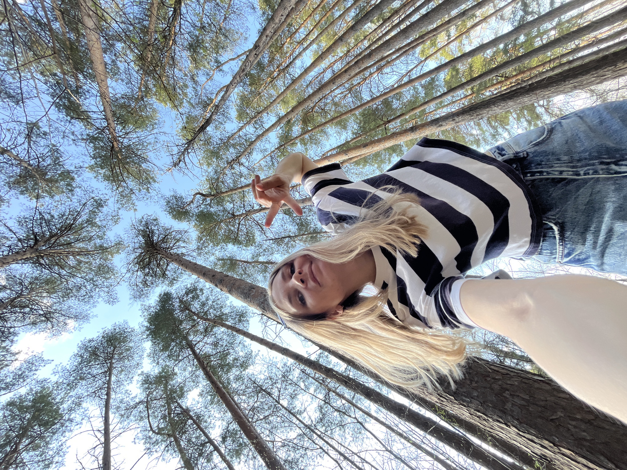 A low-angle, upward-facing photograph captures a person with blonde hair wearing a navy and white striped shirt and denim jeans, gesturing with their hand in a "peace sign" or "victory" pose. The person appears to be standing or lying down, looking directly at the camera, with a relaxed expression. The background consists of tall trees reaching towards the sky, suggesting a forest or wooded area. The sky is visible through the branches, with patches of blue and some wispy clouds. The trees' slender trunks suggest that this picture was taken in a pine forest. The location is likely in Chaikovsky, Russia, given the prompt's request for such a description.