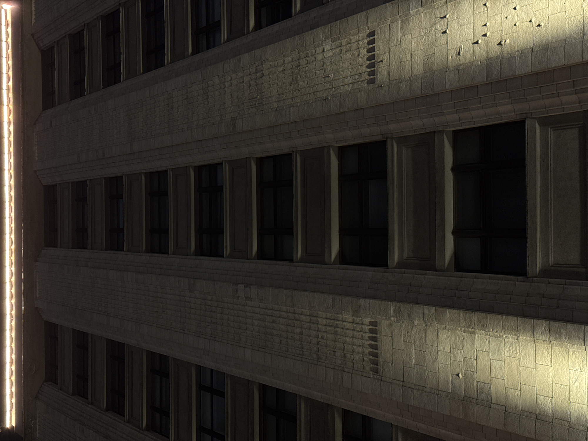 The image shows an architectural facade, likely an office building, at night. The building has rows of recessed windows. The facade is made of light-colored stone with a brick-like pattern. Some sections of the facade are illuminated with a warm yellow light, while others are shadowed. A vertical strip of light runs along the left edge of the frame, possibly a light fixture. The building's architectural style appears to be Art Deco or a similar early 20th-century design. There is no evidence to suggest this building is in Moscow, Russia.