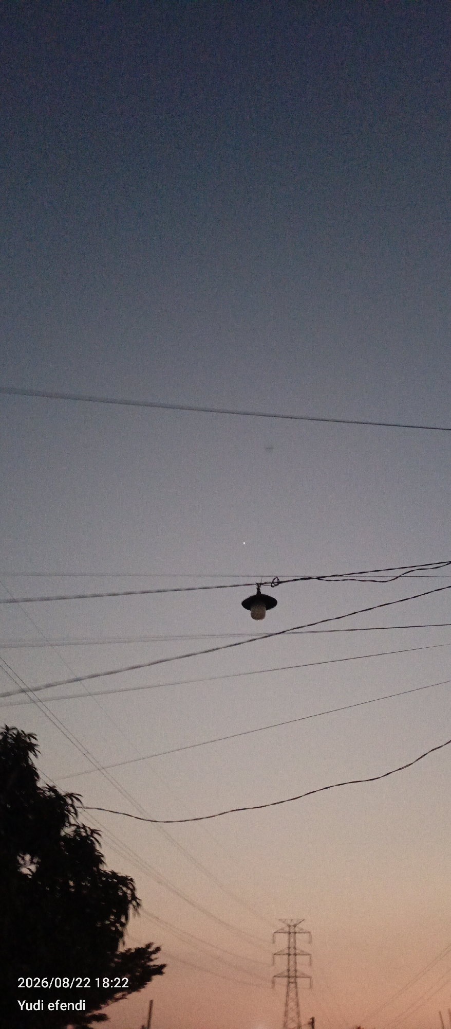 The image depicts a twilight sky over what appears to be a semi-urban or rural area. In the foreground, a silhouetted tree is visible on the left. A transmission tower, characteristic of Indonesian infrastructure, stands in the lower right quadrant, also silhouetted against the fading light. Multiple power lines crisscross the frame. A single streetlamp hangs from one of these lines, illuminated faintly. A small, bright point of light, likely a planet or a bright star, is visible in the upper portion of the sky, to the right of the streetlamp. The lower part of the sky exhibits a gradient of pastel colors, transitioning from a pale orange near the horizon to a soft grey and then a deep blue at the top. Text in the bottom left corner indicates the date and time as "2026/08/22 18:22" and a name "Yudi efendi". While the specific location of Rawajitu Selatan, Indonesia is provided, there are no visual cues within the image that explicitly confirm this location, such as recognizable landmarks, signage, or distinct architectural styles.