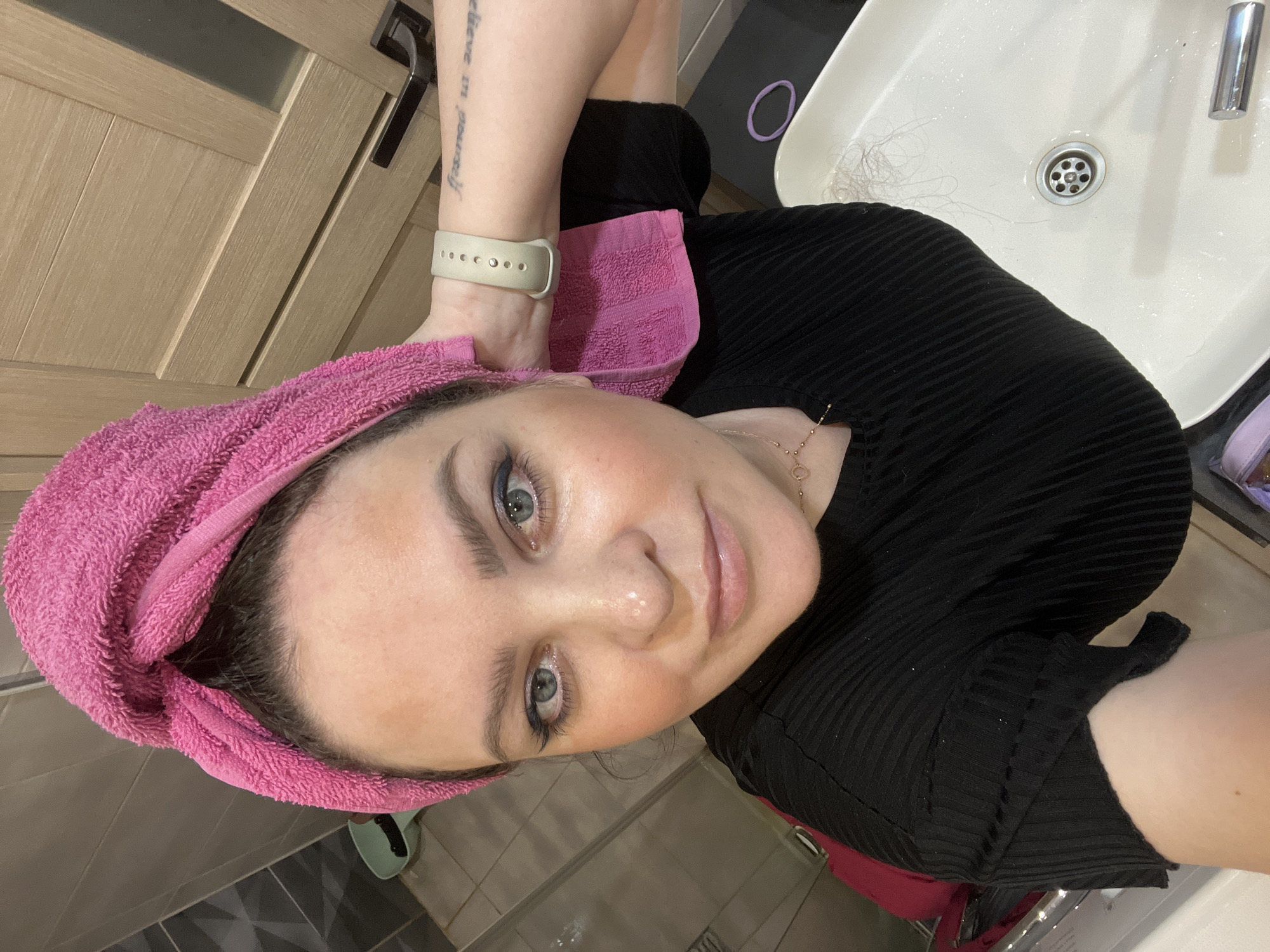 The scene is located in an indoor bathroom or dressing area in the city of Noviy, Russia. A woman, appearing to be an adult, is depicted in a close-up selfie-style photograph, looking directly at the camera with a neutral expression. Her dark hair is wrapped in a bright pink towel. Reddish-brown pigmentation or residue is visible on her forehead and cheeks. She wears a black, ribbed, short-sleeved top and a gold-colored necklace with a small heart-shaped pendant. A light beige smartwatch is on her left wrist, and a tattoo reading 'believe in yourself' or similar text is partially visible on her left forearm. Her left hand is raised, holding the pink towel in place. The background features grey-brown wall tiles with a geometric pattern on the left, and a light wooden door with a dark handle and frosted glass panel on the right. In the lower right, a white rectangular ceramic sink with a chrome drain contains several strands of hair. A purple hair tie rests on the dark countertop adjacent to the sink.