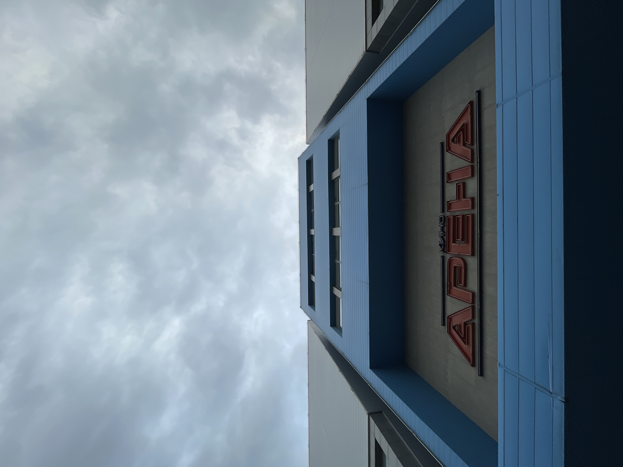 The image shows the exterior of Arena Cinema in Sofia, Bulgaria. The building has a modern design with blue and grey panels. The word "Arena" is displayed vertically in red lettering. The sky is overcast. There are no visible people or interactions.