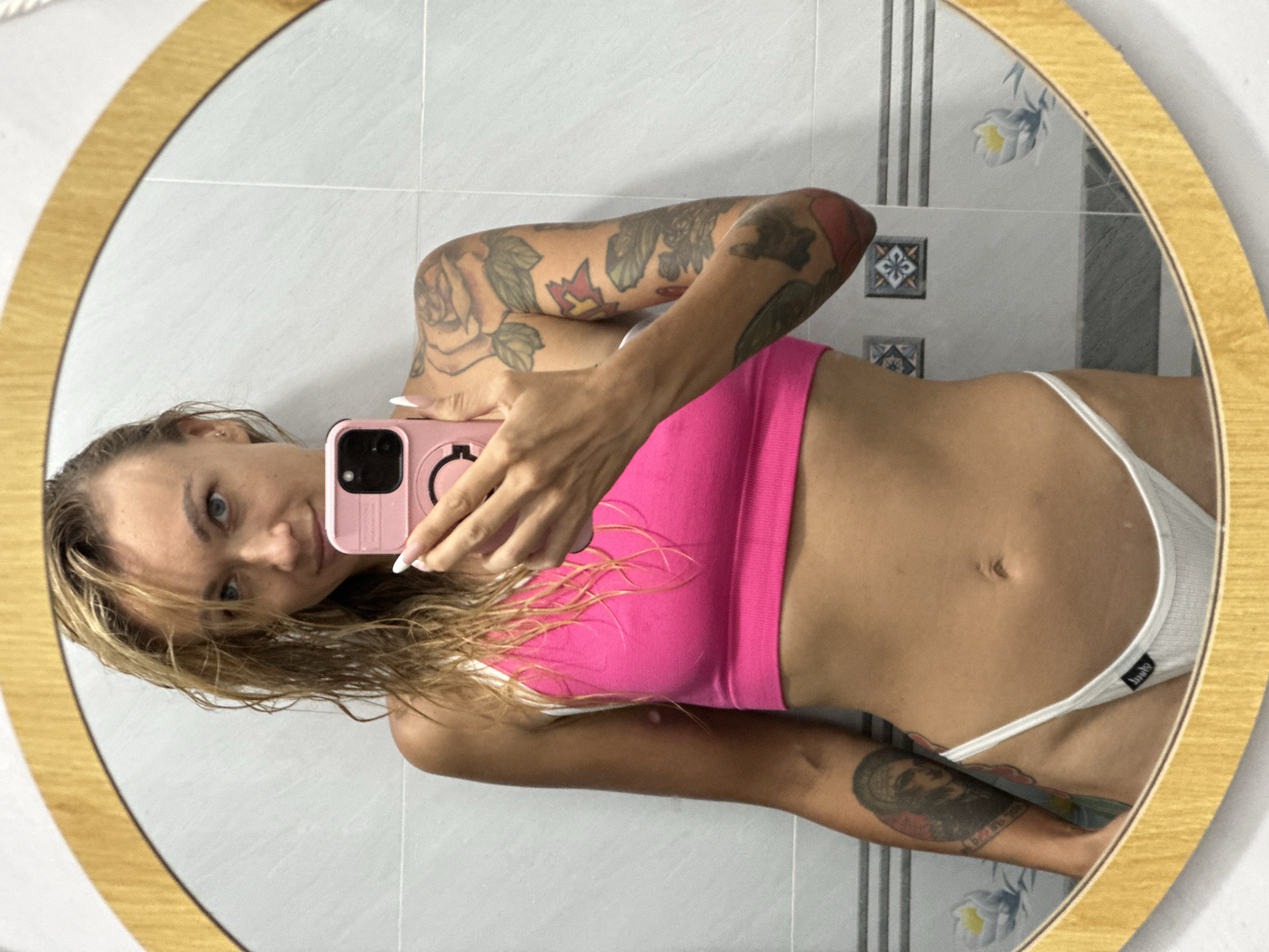 A blonde woman with blue eyes and wet hair is taking a mirror selfie. She is wearing a bright pink sports bra and white underwear. She has tattoos on both arms. The woman is standing in a bathroom with tiled walls and a gold-rimmed mirror. The reflection shows the woman holding a pink cell phone up to take the picture. The background is a tiled wall with decorative elements.