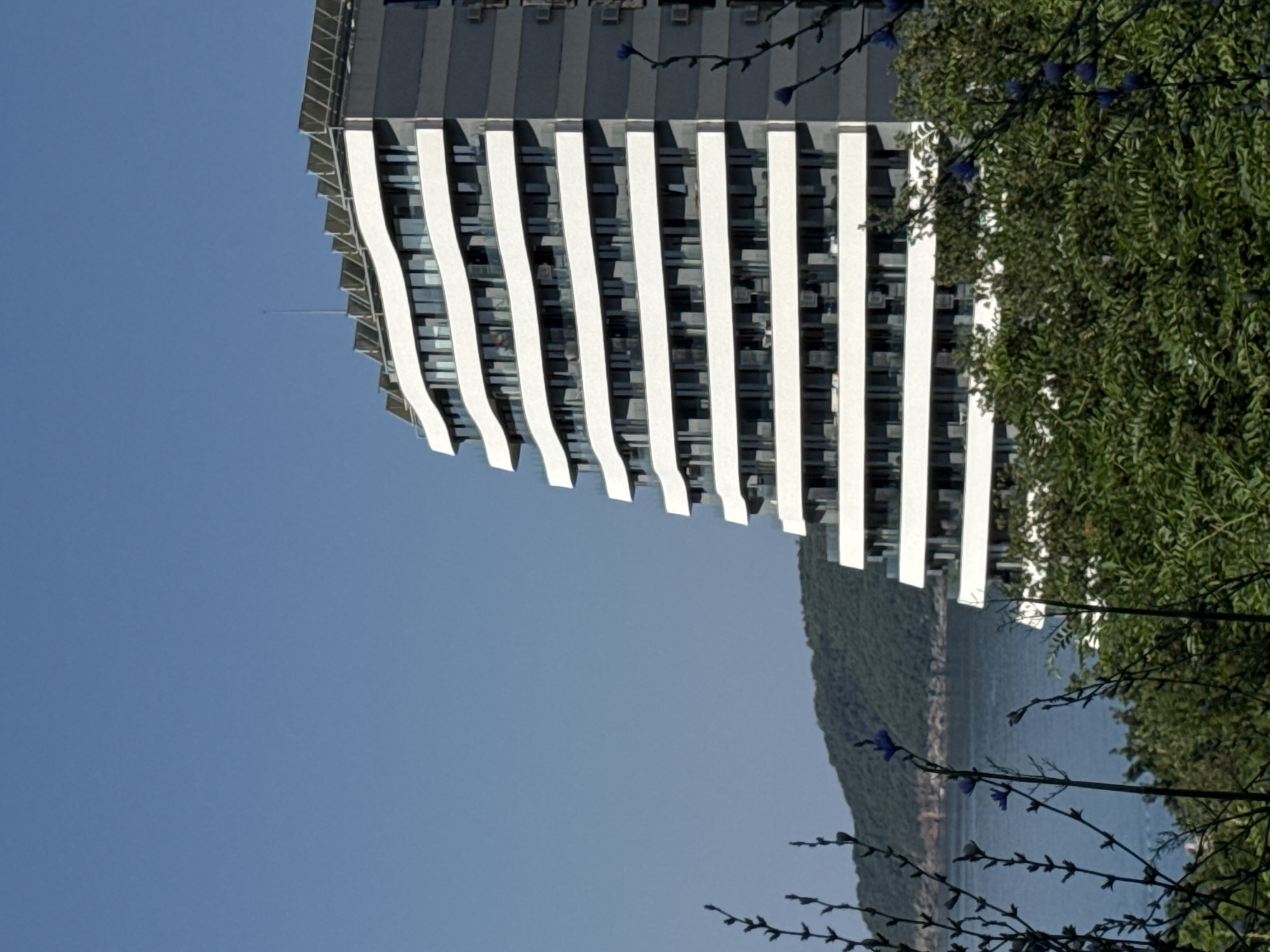 This media file showcases a section of a modern building with a distinctive facade. The building features a series of white, vertical elements that curve outwards at the bottom, creating a visually dynamic effect. Behind these white elements, windows are visible, suggesting multiple floors. The building's upper portion has a darker, more solid appearance.
To the right of the building, lush green foliage is present, with some delicate branches and small purple flowers extending into the foreground. The foliage creates a natural contrast to the man-made structure.
In the background, a body of water, likely the sea, is visible, bordered by a densely vegetated hillside. The sky above is a clear, bright blue, indicating a sunny day with fair weather. The scene appears to be set in a coastal or waterside location, given the presence of the water and the hilly landscape.
The orientation of the image is such that the building runs diagonally from the top left to the bottom right. There is no visible text in the image. The overall impression is one of a harmonious blend between modern architecture and the natural environment.