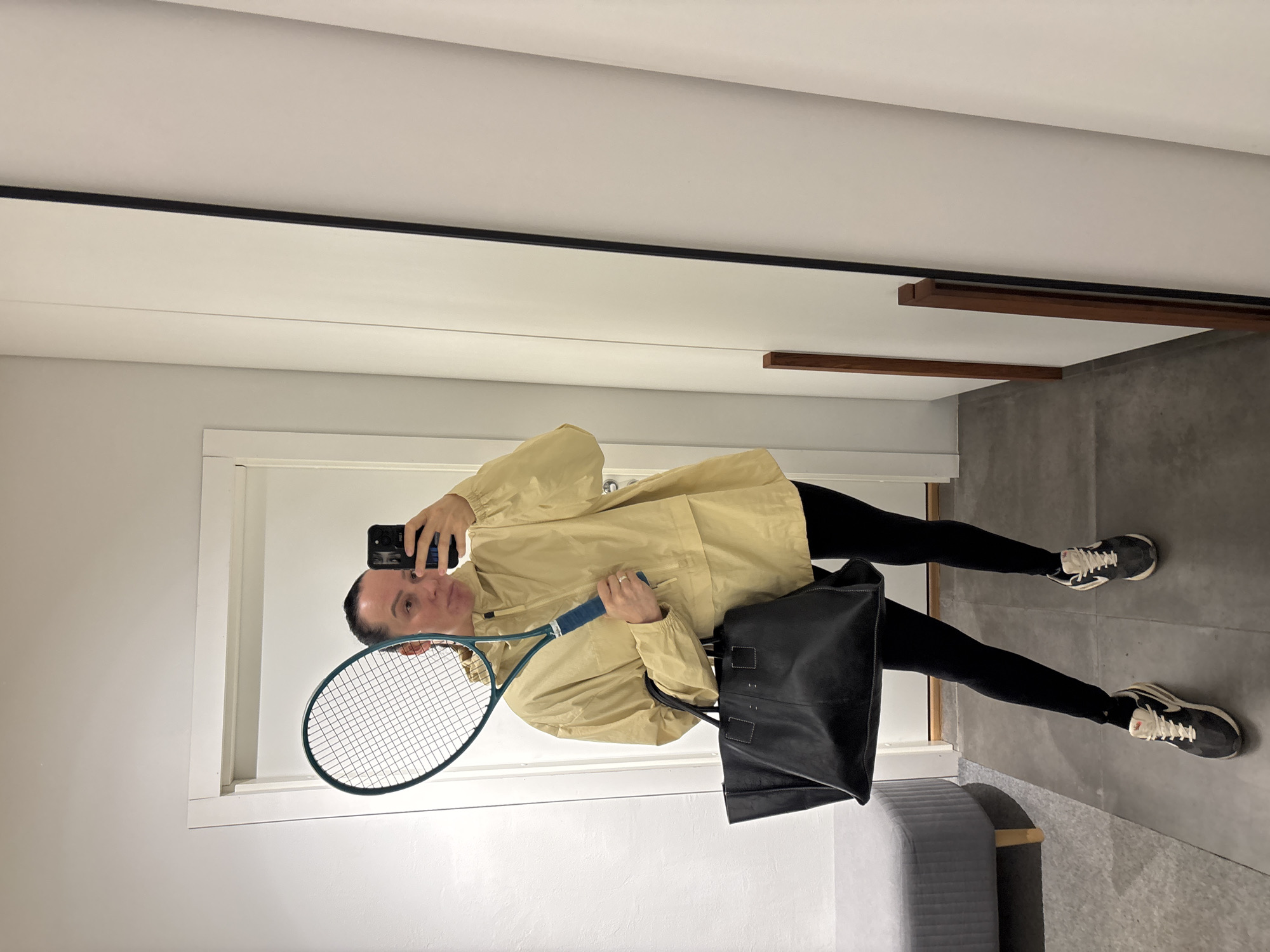 A person is taking a selfie in a mirror. The person is holding a tennis racket in their left hand and a black bag in their right hand. They are wearing a light yellow jacket, black leggings, and black and white sneakers. The person's hair is dark and pulled back. The background includes a white door, a gray floor, and a gray bench. The location is not identifiable as the city of Chaikovsky, Russia.