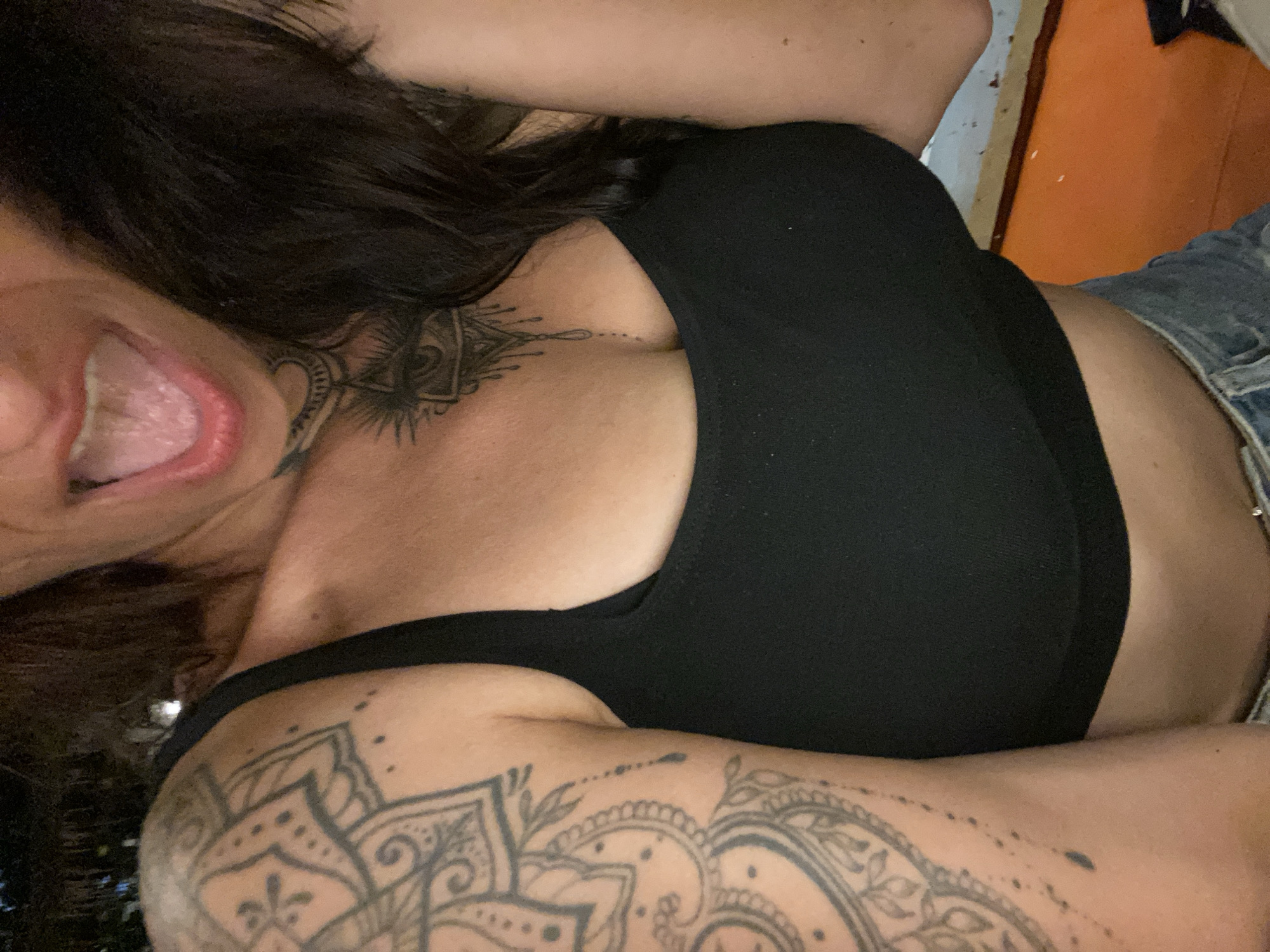 A close-up, waist-up shot shows a woman with dark hair, a black tank top, and blue jeans. She has extensive tattoos on her left arm, chest, and neck. Her mouth is open, with her tongue visible. The background is dark and out of focus, with hints of orange and blue. The scene is likely an outdoor setting at night, possibly in a public area.
