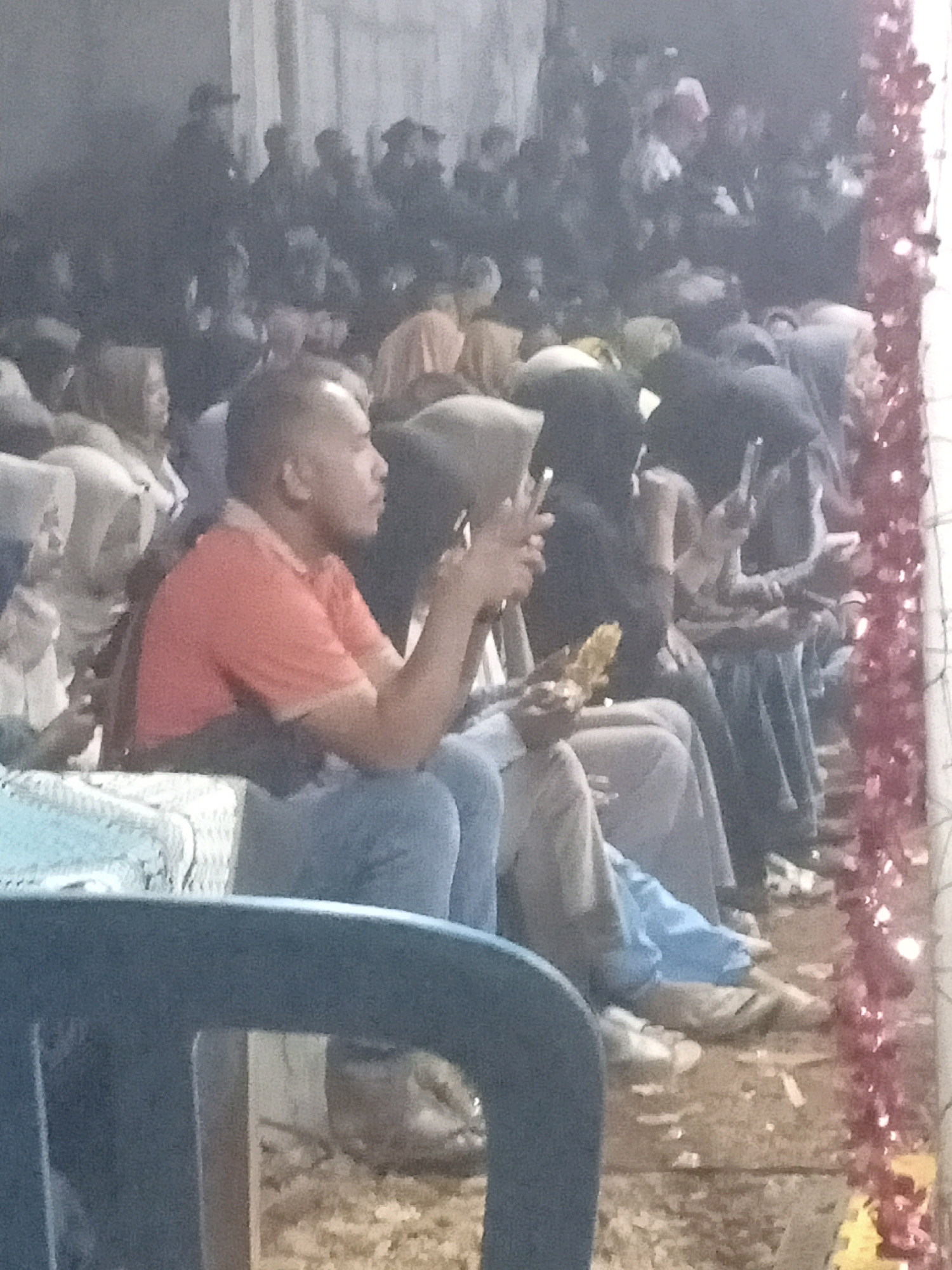 In Tasikhargo, Indonesia, a group of people are gathered, some seated in chairs, while others are standing or sitting on the ground. A man in an orange shirt is holding a phone and appears to be looking at it. To his right, a woman in a hijab is also holding a phone. In the background, a crowd of people are visible, some of whom are also holding phones. The scene appears to be an outdoor event, possibly a festival or gathering, given the presence of decorative lights on the right side of the image. The ground is covered with dirt and debris. The lighting is dim, suggesting it may be evening or nighttime.