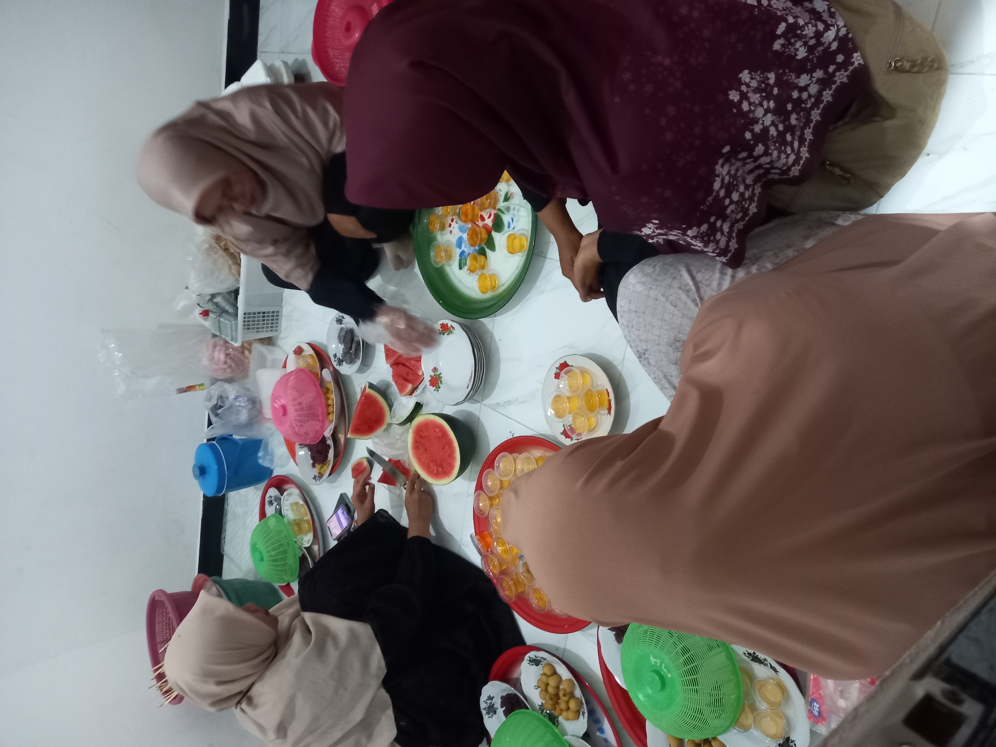 Four women, all wearing hijabs, are sitting on the floor in a room, likely in Tempos, Indonesia, gathered around plates of food and drinks. One woman, in a light brown hijab and black top, is holding a knife and cutting a slice of watermelon. Another woman, in a light brown hijab and tan pants, is seated to her right, looking down at her hands. A third woman, in a maroon hijab with white floral patterns and a tan skirt, is seated in the foreground, with her hands clasped. The fourth woman, in a light beige hijab and black top, is seated on the far left, looking towards the center.
The floor is tiled with a white and gray marble pattern. Several red and green plastic trays are on the floor, holding various items. One tray contains small cups filled with yellow liquid, likely a dessert or drink. Another tray holds longan fruit. There are also plates of watermelon slices and dark, possibly dried, fruits. In the background, there are plastic bags, a blue plastic bucket, and two pink round containers. The overall atmosphere suggests a casual gathering or meal.
