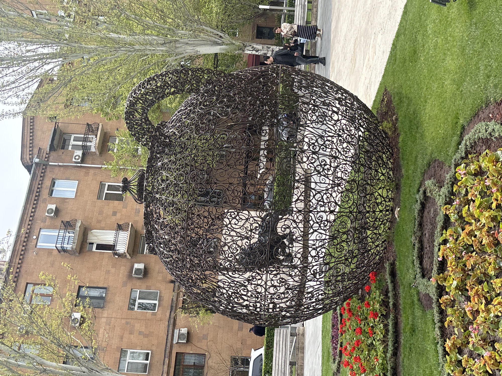 The image depicts a large, ornate, metal sculpture in the shape of a teapot. The sculpture is positioned in a well-manicured park area with grass, flower beds containing red and yellow flowers, and a paved walkway. In the background, there is a multi-story brick building with balconies and windows. Several people are walking along the walkway in the background. The setting appears to be an urban park in Yerevan, Armenia, given the architectural style of the building and the presence of the teapot sculpture, a known landmark in the city.