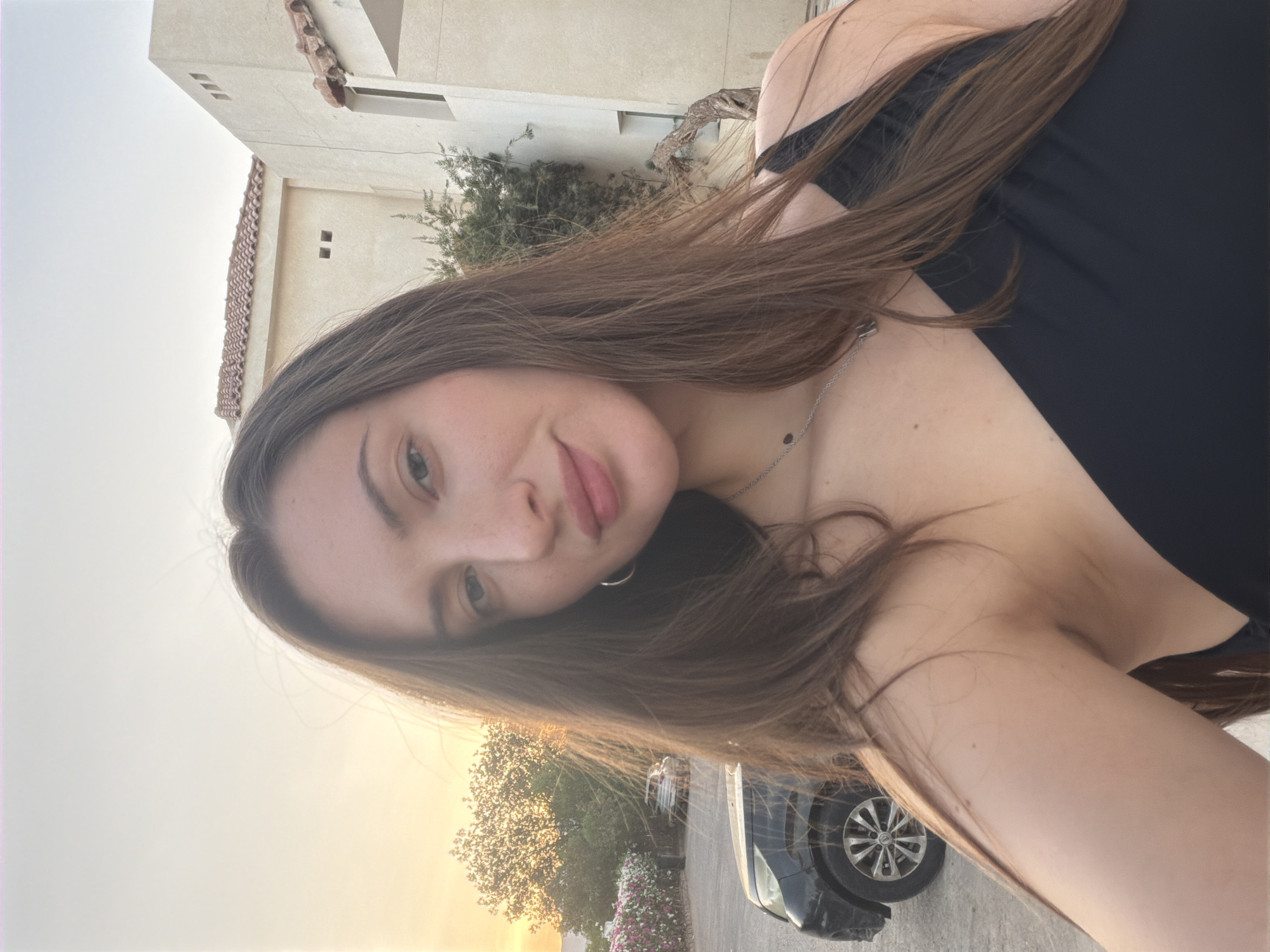 A young Caucasian woman with long brown hair is taking a selfie outdoors, likely in Sharm El Sheikh, Egypt, as evidenced by the distinct architecture and landscaping. The setting appears to be near a residential or resort area, with a multi-story building featuring a tiled roof and stucco exterior visible in the background. There's also a dark-colored car parked to the left, and lush vegetation with pink flowers suggesting a warm climate. The woman is wearing a black, off-the-shoulder top and a delicate silver necklace. The sun appears to be setting, casting a warm glow over the scene, and the sky is a soft, pale orange. Her expression is neutral, with her head tilted slightly.