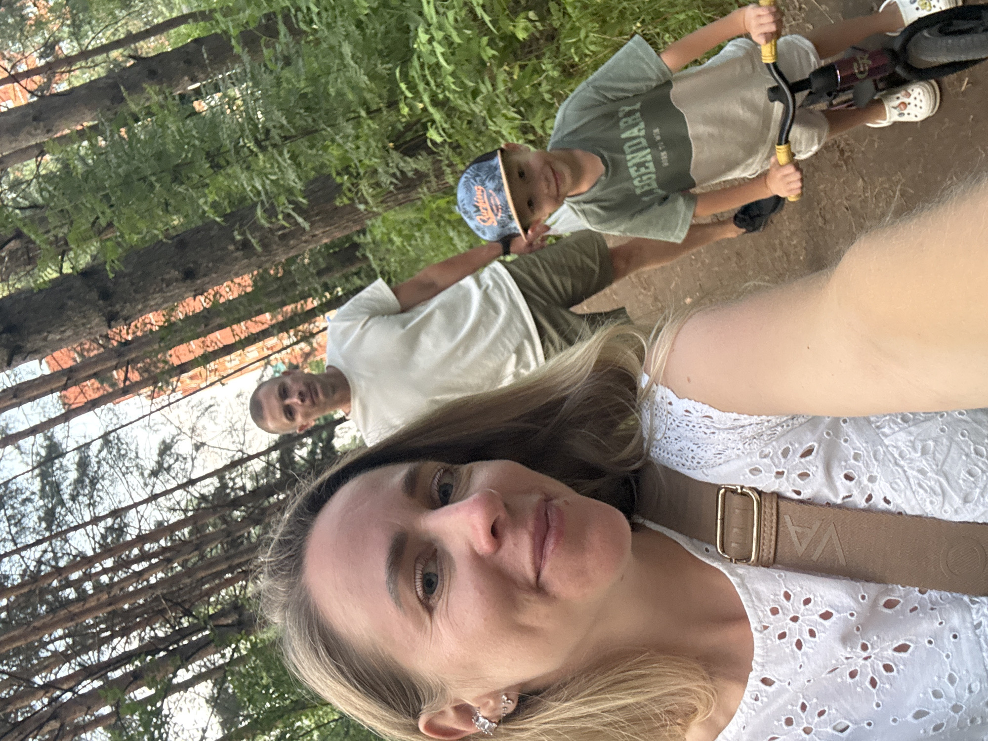 In the city of Chaikovsky, Russia, a woman is taking a selfie with her family in a park. She is in the foreground, smiling at the camera. Behind her, a man is walking, and a young boy is riding a balance bike. The family is surrounded by trees and lush greenery. The sun is setting, casting a warm glow over the scene.