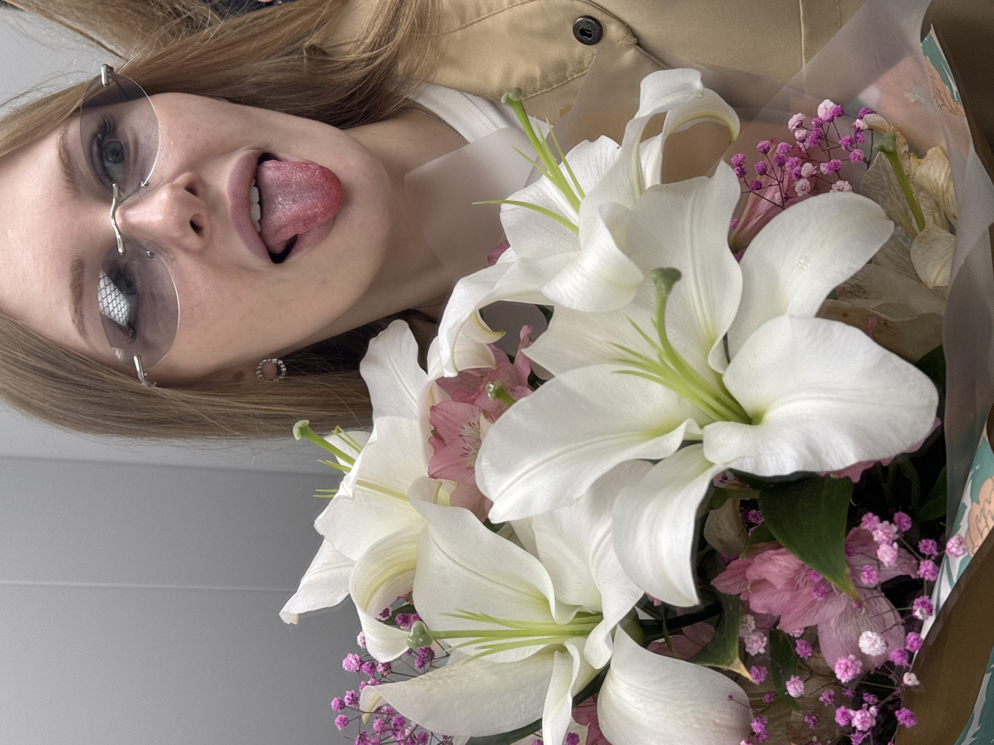 The image shows a young Caucasian woman holding a bouquet of white lilies and pink flowers. She is wearing rimless sunglasses and has her tongue sticking out. The background appears to be an indoor setting with gray walls. The image does not provide any information about the city of Perm, Russia.