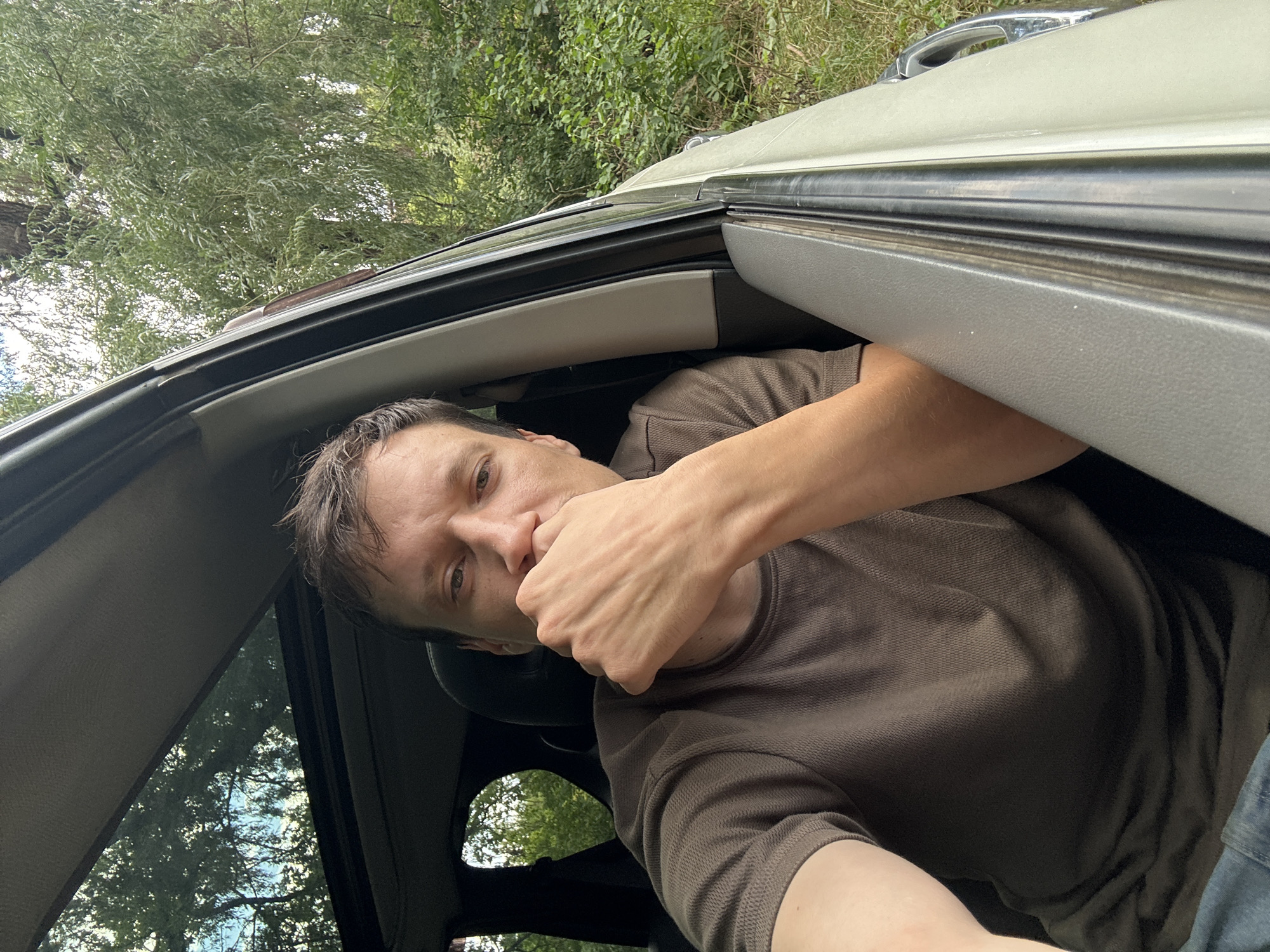 In an exterior, medium shot, a man is seated inside a vehicle, his head and upper torso visible through the open window. He has short, brown hair and is wearing a brown, short-sleeved shirt and blue jeans. His right hand is raised to his face, with his fingers touching his mouth and chin. The man's gaze is directed towards the camera. Through the window and the vehicle's sunroof, trees and foliage are visible, suggesting an outdoor environment. The background is predominantly green, indicating a natural setting. No specific landmarks of Makiv, Ukraine, are identifiable.