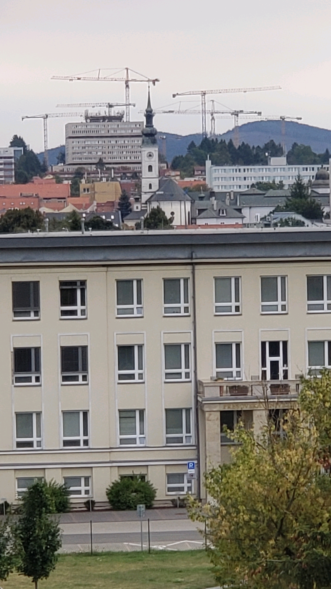 The image depicts an urban landscape in Prešov, Slovakia, dominated by the facade of a light-colored, multi-story building with numerous windows. In the background, a cityscape unfolds, characterized by various buildings, including a prominent church with a spire and a taller, older building topped with construction cranes. The sky is overcast, suggesting a cloudy day. The foreground shows a grassy area with trees and a fence, indicating a park or a residential area adjacent to the building. A parking sign is visible on the side of the building.