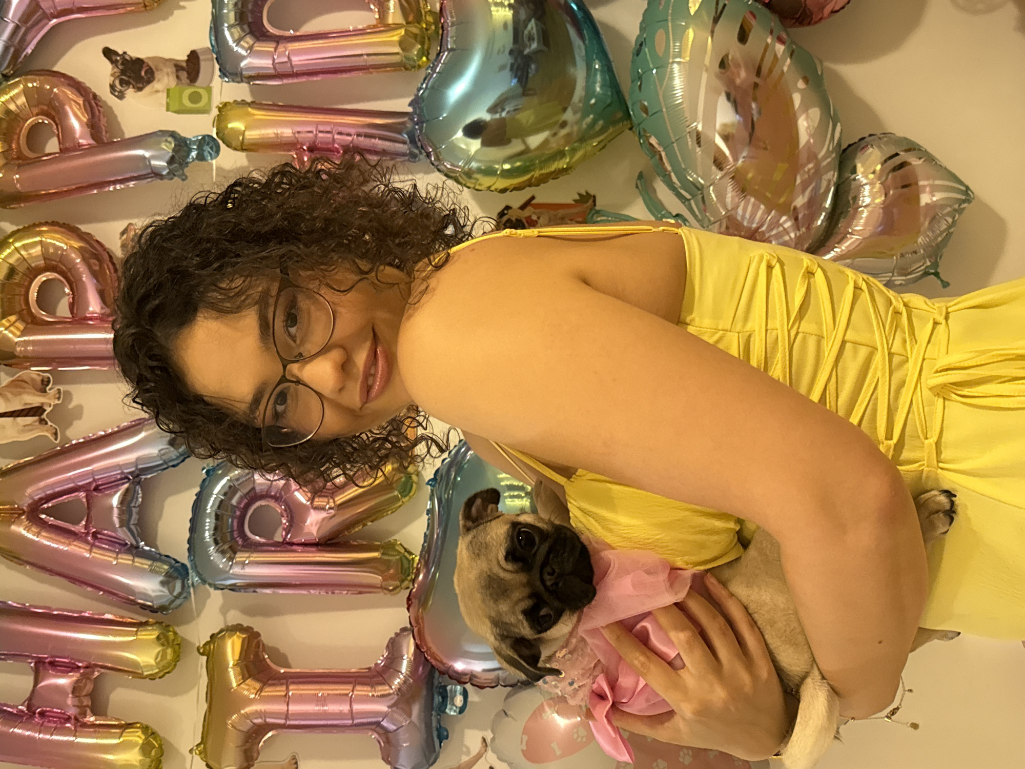 An image, rotated 90 degrees counter-clockwise, depicts a young woman holding a pug in front of a wall decorated with numerous foil balloons.
The primary subject is a young woman with curly brown hair and round glasses, smiling warmly at the camera. She is wearing a bright yellow, possibly strapless or thin-strapped top or dress. She holds a small, fawn-colored pug with a black mask, which is dressed in a pink, ruffled outfit. The pug's head is tilted slightly, and its large, dark eyes look directly at the viewer. The woman's hands are gently supporting the dog, showcasing affection.
The background is a light-colored wall covered with an assortment of metallic, iridescent foil balloons in shades of pink, blue, gold, and silver. Many of these balloons are shaped like letters, suggesting a celebratory message. Visible letters include parts of 'H', 'A', 'P', 'P', 'Y', 'B', 'I', 'R', 'T', implying words like "HAPPY BIRTHDAY" or a similar festive greeting. There are also heart-shaped balloons and possibly other decorative shapes. The overall scene conveys a cheerful and celebratory atmosphere, likely a birthday party or a similar special event. The lighting is even, suggesting an indoor setting.