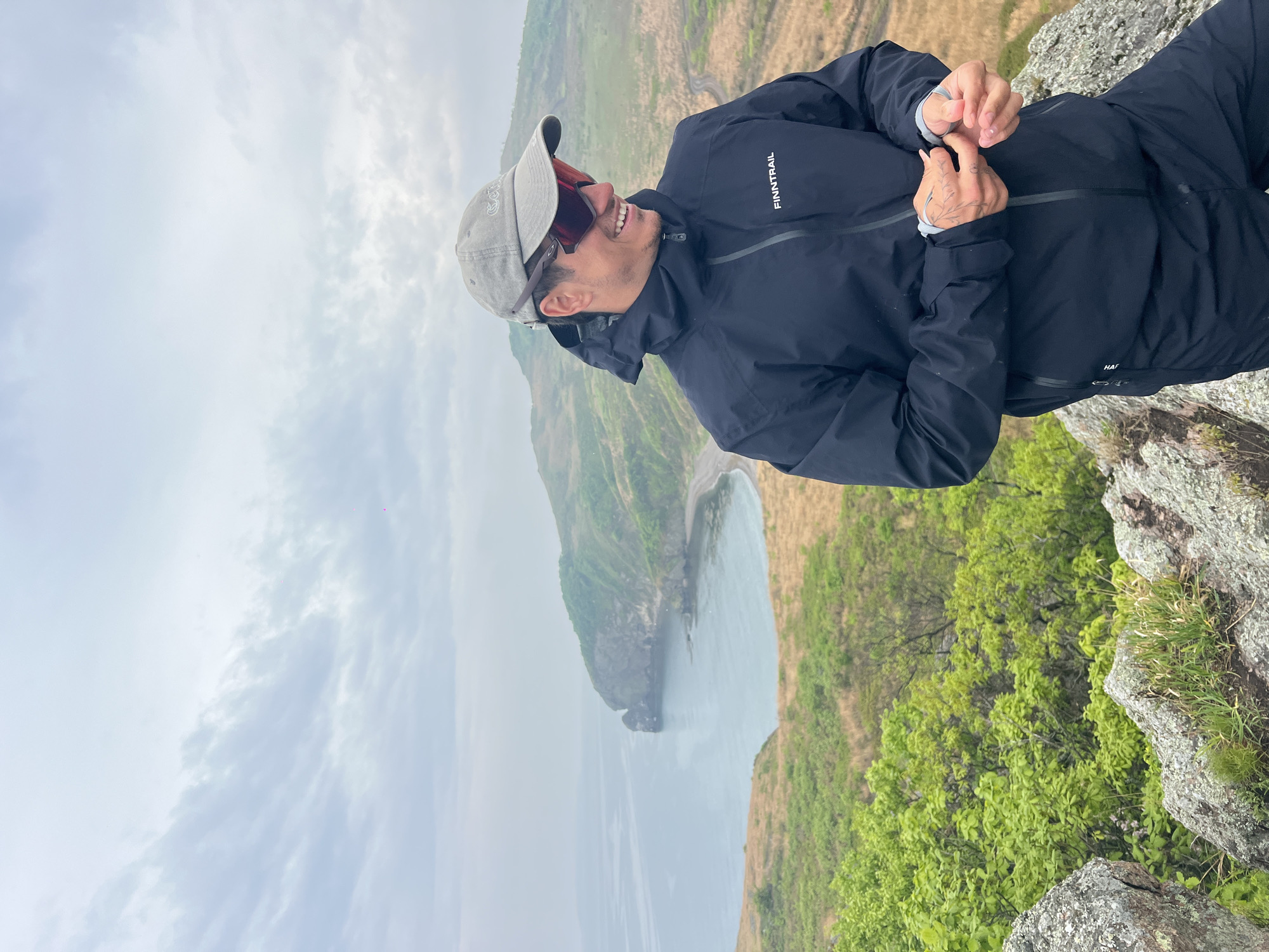 A medium shot depicts a man standing on a rocky precipice overlooking a bay. The man is clad in a black jacket, a grey cap, and dark sunglasses. He is smiling and looking to his left. His hands are raised near his chest. The jacket has the word "FINNTRAIL" on the upper left chest. In the background, a body of water stretches towards the horizon under a cloudy sky. To the left, a green, sloping cliff extends into the water. To the right of the man, there is green vegetation growing on the rocky terrain. The location is Gvozdevo, Russia.