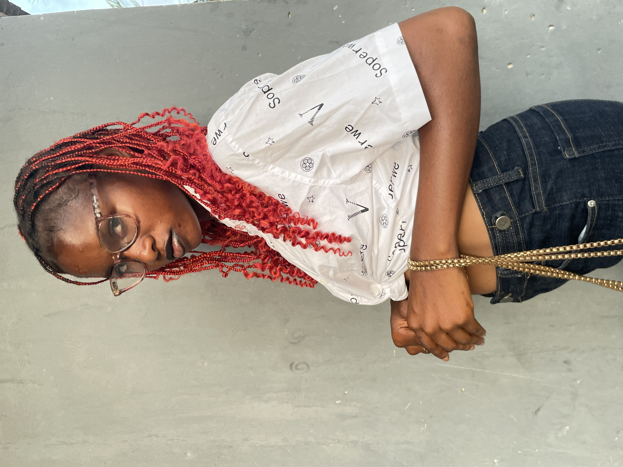 The image, taken in Badagry, Nigeria, features a young Black woman positioned against a plain, light grey background. She is captured from the chest upwards, with the image rotated 90 degrees clockwise. She has striking red braided hair, styled in numerous small twists.
The woman wears round, light-rimmed eyeglasses and gazes towards the viewer with a neutral, slightly pensive expression, her mouth subtly parted. She is dressed in a white collared shirt adorned with repetitive black patterns, including stars, soccer ball symbols, and stylized text. Visible text on her shirt includes "Soperwe" (or "Superwe") and the mirrored letters "ewr". Below her shirt, a portion of dark blue denim shorts or jeans is visible.
A prominent detail is that her hands are brought together in front of her at the wrists, and she appears to be bound or tied by multiple strands of a gold-colored chain or rope. The lighting is bright and even, consistent with daylight, though no specific time or weather is discernible. The background is simple and unadorned, providing no additional scene cues beyond its uniform grey surface, possibly a wall or floor.