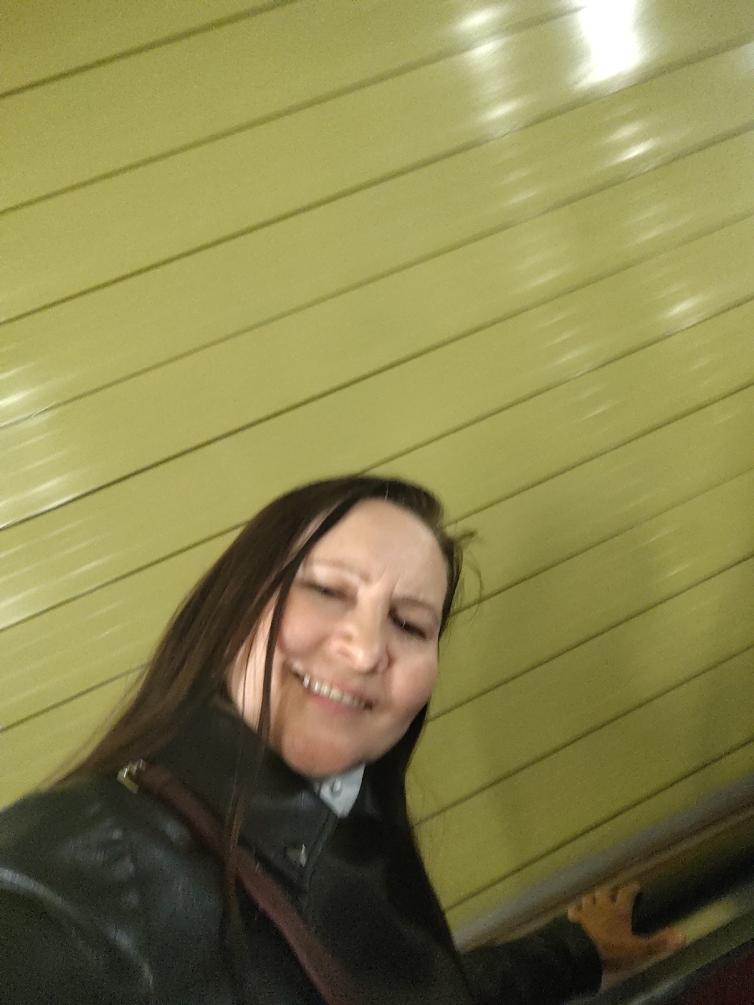 A woman is looking at the camera and smiling. She is wearing a dark-colored jacket and standing next to what appears to be a moving walkway or escalator, with a paneled wall visible in the background. The location and city are unknown.