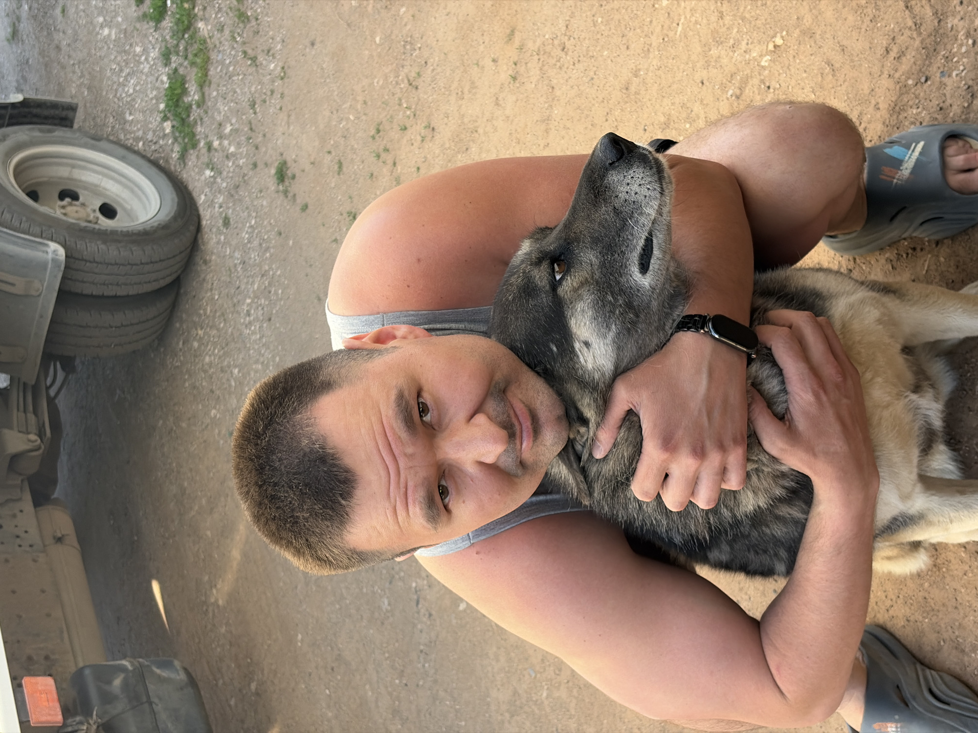 A man with short, dark hair and fair skin is hugging a large dog. The man is wearing a gray tank top and a black smart watch on his left wrist. He has on gray Crocs on his feet. The dog is mostly gray and tan, with a black muzzle. The man is holding the dog tightly. The background appears to be a dirt or gravel surface. A truck wheel and part of the truck are visible on the left. The location is unidentifiable as Izhevsk, Russia.