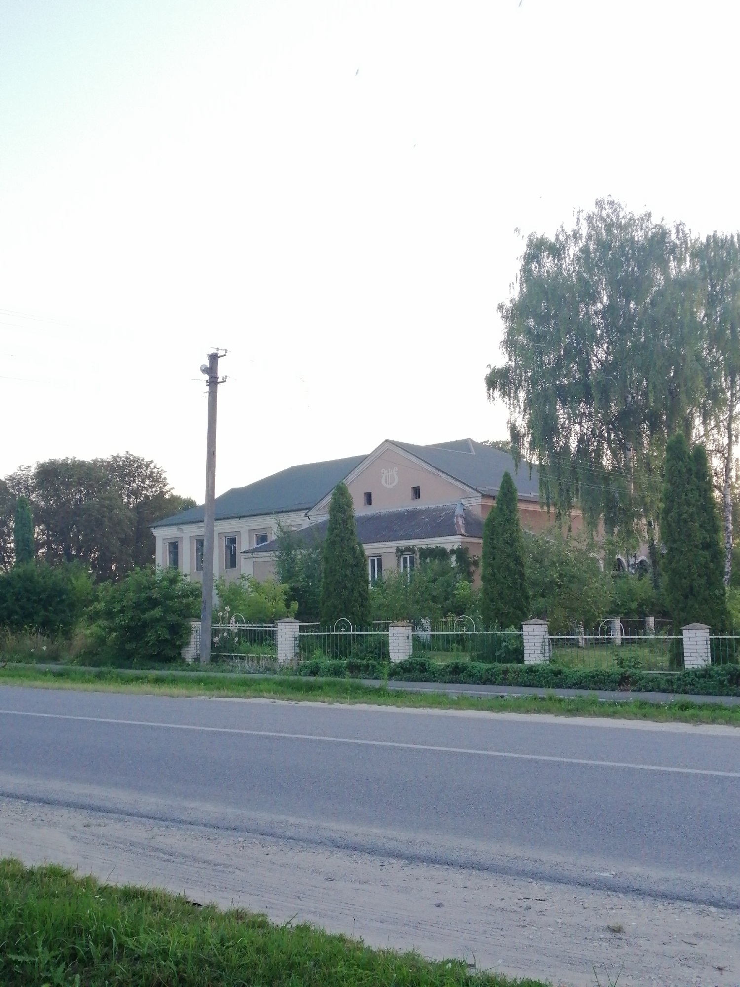 The image shows a two-story building with a light-colored facade and a dark roof, likely a public institution or residence, situated in a rural or suburban area. A fence made of brick posts and metal bars runs in front of the building, separating it from a paved road. The building has several windows and an emblem above the entrance. Tall green trees and bushes surround the property. A utility pole with wires stands to the left of the building, and a paved road with a white line marking the lane runs across the foreground. The sky is overcast and pale. The location is identified as the city of Lanivtsi, Ukraine. No people are visible in the image.