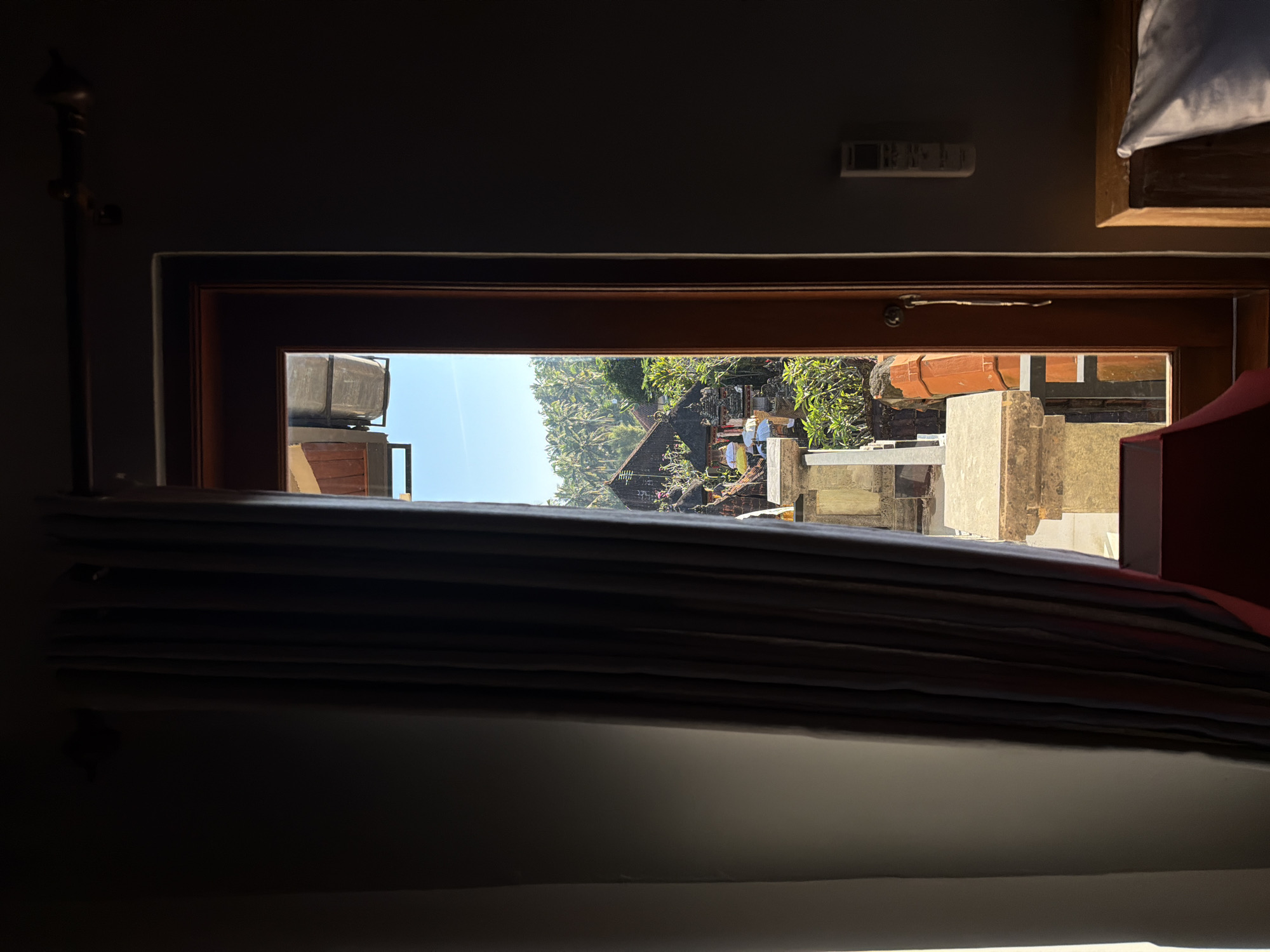 The image shows a view through a doorway in Tegallalang, Indonesia. On the left, a dark gray curtain is partially drawn, revealing a glimpse of a room interior. The doorway itself is framed in dark wood and opens to an outdoor scene bathed in bright sunlight.
Outside, a stone balustrade or railing is visible, topped with green plants. Beyond this, several traditional Balinese structures with dark roofs are nestled among lush green foliage and palm trees under a clear blue sky. White umbrellas are dotted around the scene, suggesting an outdoor gathering or ceremonial area.
To the right of the doorway, the wall of the interior room is visible, painted a dark gray. A white air conditioning unit is mounted on this wall. In the foreground on the right, a wooden bed frame with a white pillow is partially visible. The overall impression is a serene view of a tropical landscape from a comfortable interior space.
