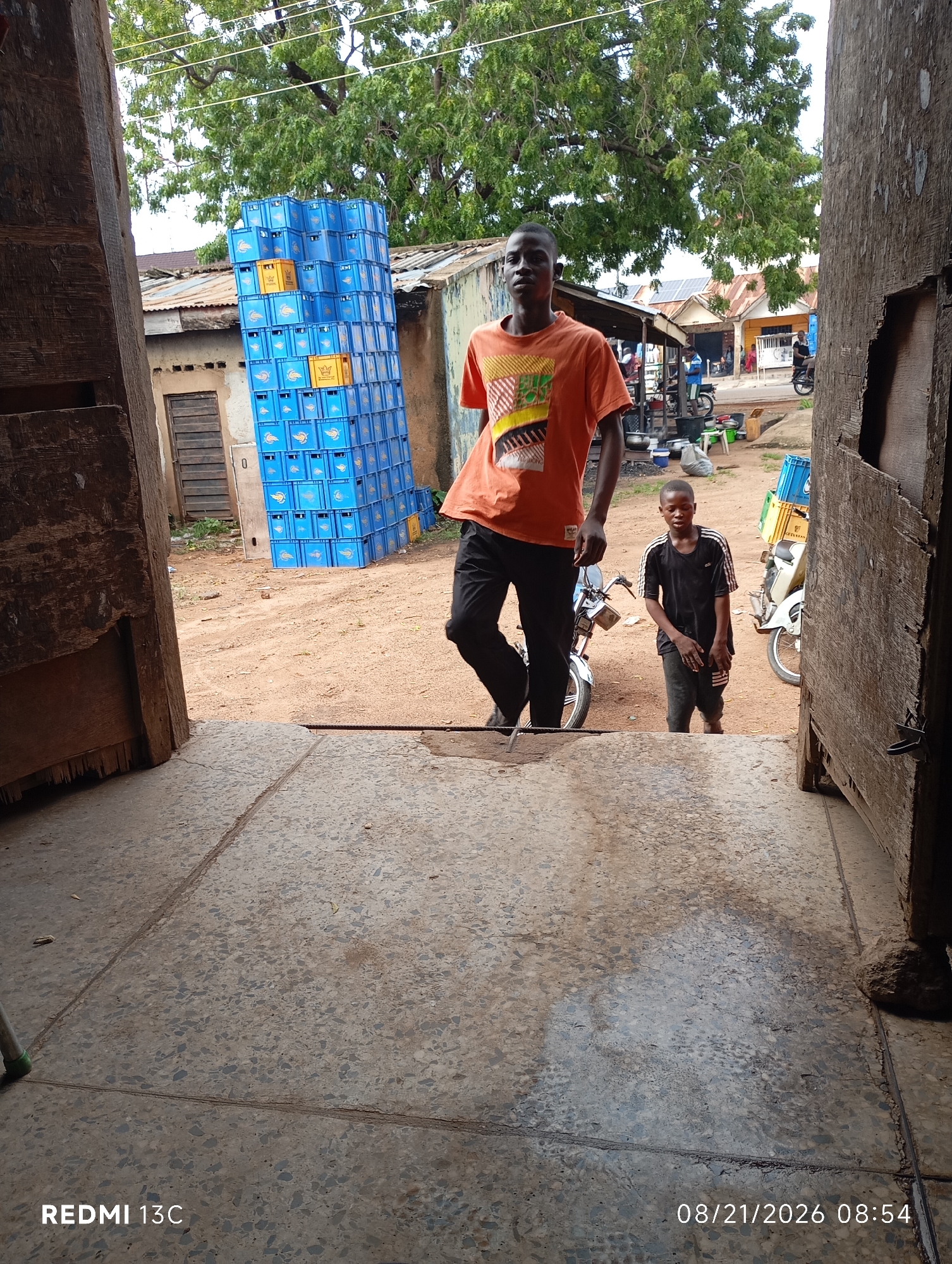 In Shendam, Nigeria, two young males are observed standing in an outdoor environment. The individual on the left is wearing an orange t-shirt with a graphic design and dark pants. The other male, positioned to the right, is dressed in a black t-shirt with white stripes and dark pants. Behind them, a large stack of blue plastic crates is visible, suggesting a commercial or storage area. The ground appears to be dirt and gravel. The scene is partially framed by wooden structures.