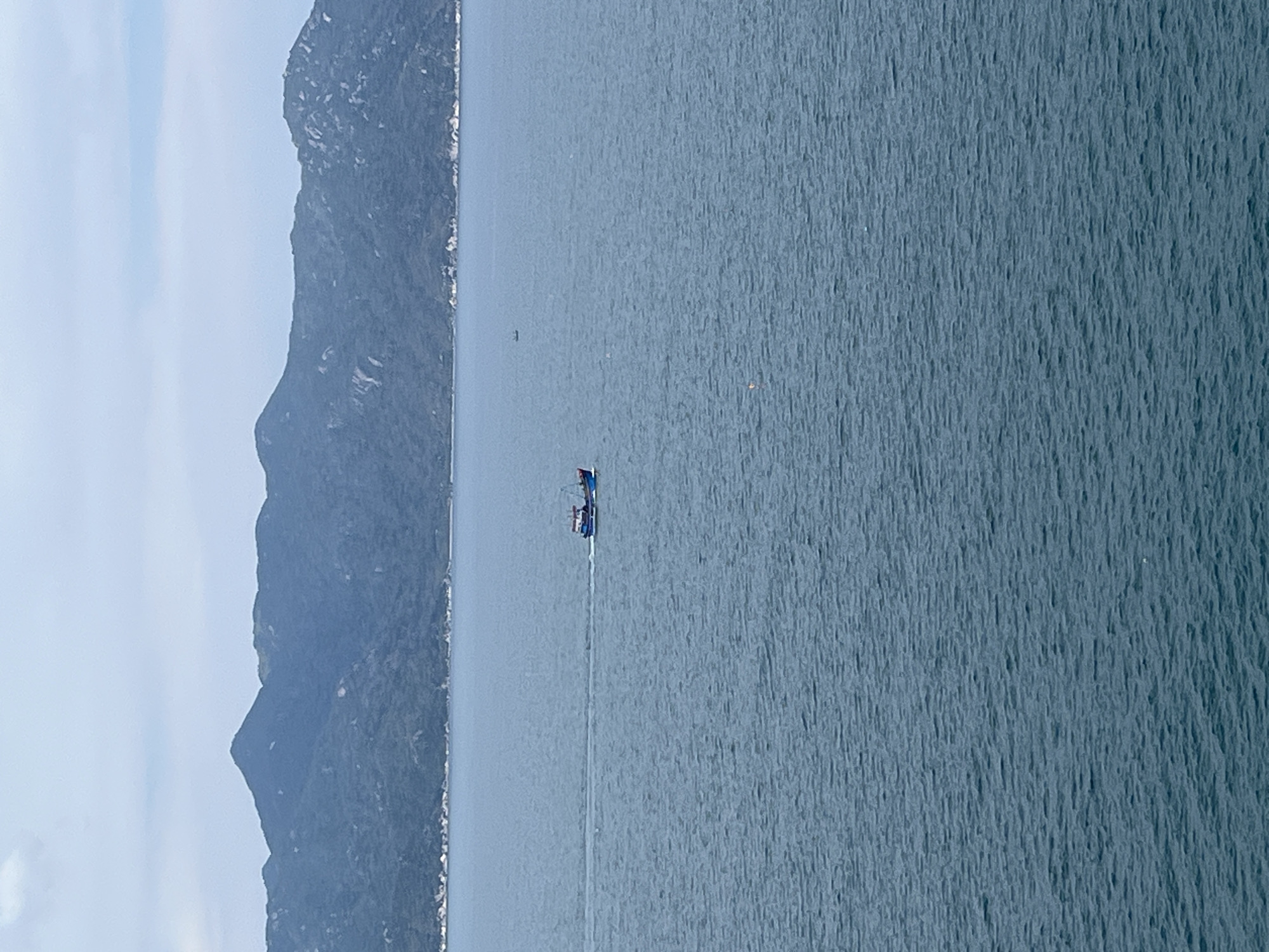 The image shows a body of water, likely a bay or sea, next to a mountainous region on the left side of the frame. In the middle of the water, there is a small boat. The water is relatively calm, and the sky is partially cloudy. This location is at Bac Nha Trang Ward, Vietnam.