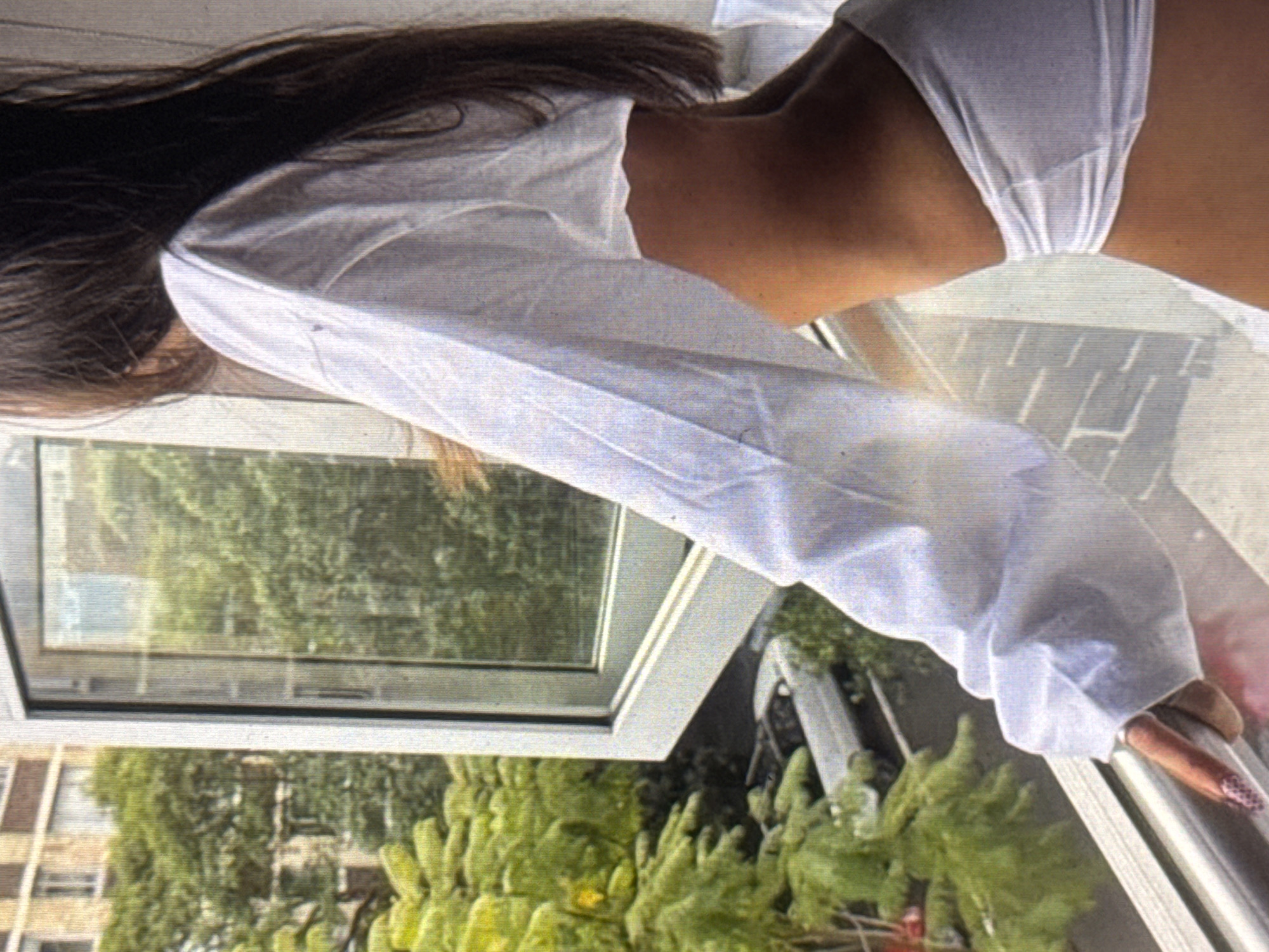 The media file shows a close-up of a person leaning out of a window. The person has long, dark hair and is wearing a white shirt and white bottoms. Their back and arm are visible, as well as part of their legs and a hand with patterned nail polish resting on the windowsill. The setting appears to be outdoors, with greenery and buildings visible through the open window. The overall lighting suggests it is daytime. The scene has a relaxed and casual feel.