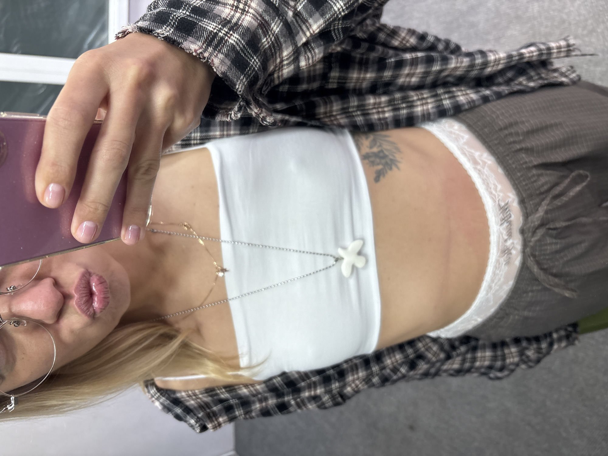A woman, wearing glasses, a white tube top, a plaid shirt, and grey sweatpants, takes a selfie with a pink phone. She has blonde hair and is puckering her lips. A tattoo is visible on her hip. The background appears to be an indoor setting, possibly a room, with a gray floor. There is no discernible connection to Ust-Ilimsk, Russia, in the image.