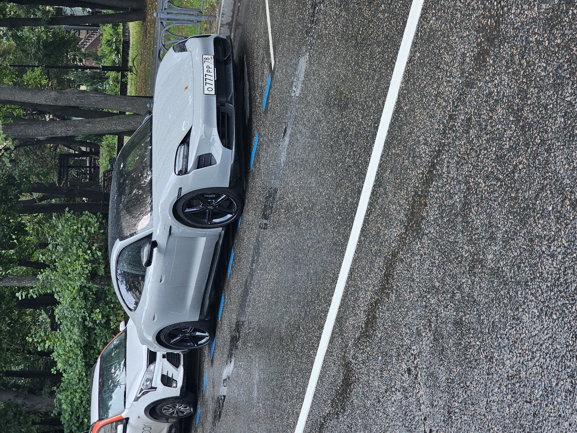 The image captures a street scene in Moscow, Russia, likely in a park or residential area, on a rainy day. The asphalt road is wet, with visible puddles reflecting the surroundings. In the foreground, a white line marks a parking space.
Dominating the mid-ground are two parked cars. The car on the left is a white compact SUV with an orange stripe on its roof and the text "car" on its side, suggesting it might be a shared mobility vehicle. This car is parked slightly angled.
To its right, a sleek, light gray Porsche Taycan is parked parallel to the curb. This luxury electric car has black rims and a black roof. Its license plate is visible and reads "O777PP 78", indicating it is registered in St. Petersburg (region 78). Blue markings are visible on the asphalt in front of the wheels of both cars, likely indicating designated parking spots, possibly for electric vehicles.
The background features a lush green environment with mature trees and dense foliage, typical of a park or a tree-lined street. A metal fence runs along the edge of the street, separating it from the park area. A park bench and lampposts are visible in the distance, further suggesting a park or a well-maintained urban green space. The overall atmosphere is subdued due to the overcast weather and rain. There are no people visible in the image.