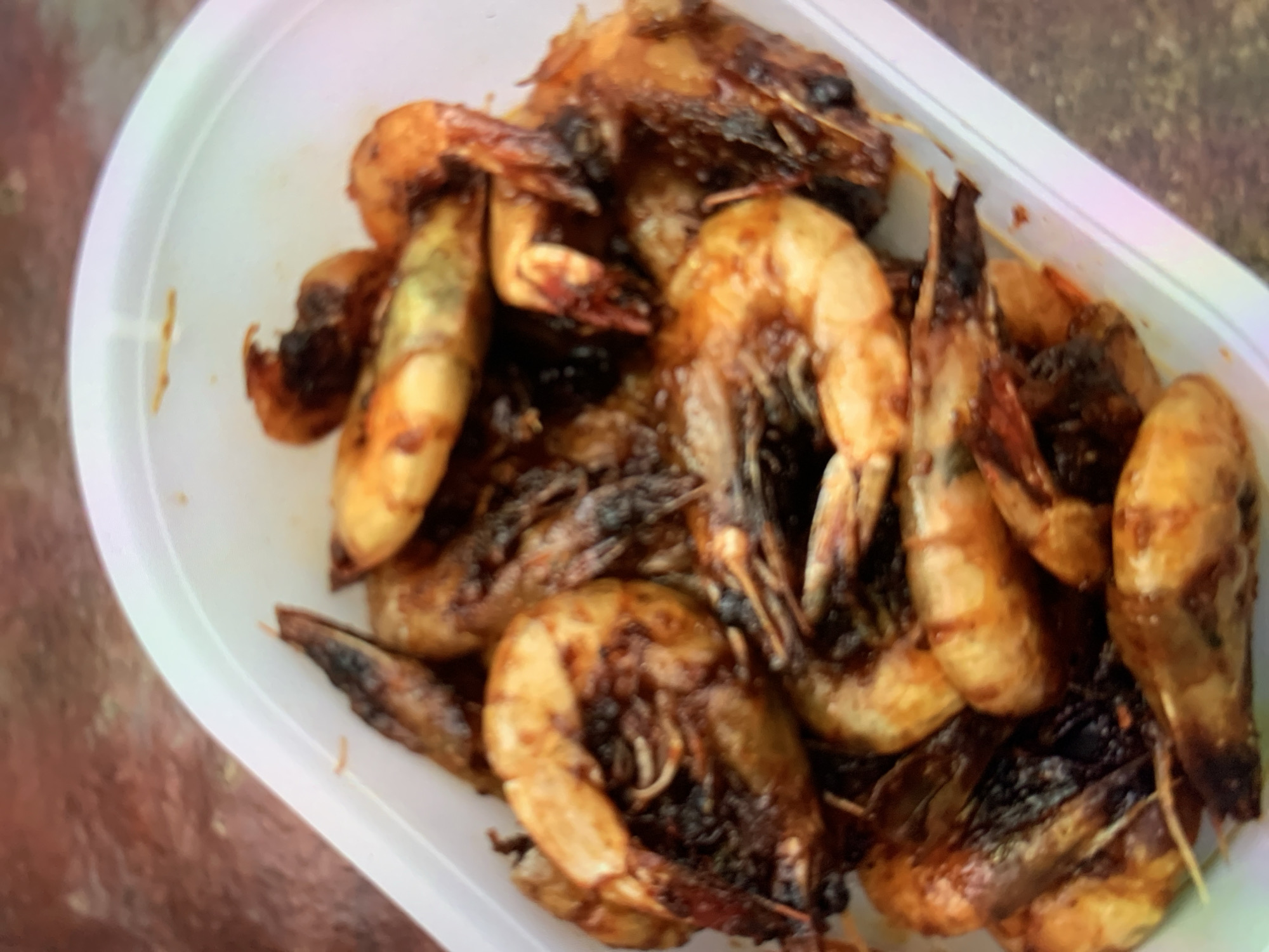 A plastic container holds a generous portion of cooked shrimp, most of which appear to be seasoned and grilled, exhibiting a dark, slightly charred appearance. The shrimp are arranged in a jumbled pile within the white container. The background is blurred, showing a textured, reddish-brown surface. This image does not contain enough information to determine a specific location within Ciamis, Indonesia.