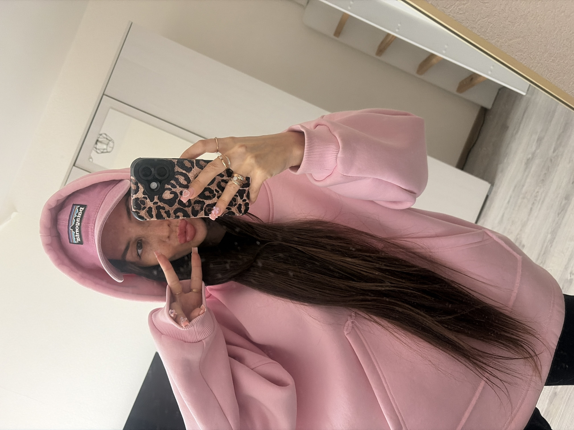 A young woman with long, dark hair and light skin takes a mirror selfie indoors. She is wearing a pink hooded sweatshirt and a pink baseball cap with a Patagonia logo. She is holding her phone, which has a leopard print case, in her right hand and is making a peace sign with her left hand. Her lips are pursed. Her nails are painted a light pink with floral accents. She is wearing several rings on her fingers. The background shows a mirror reflecting a room with white furniture and a wooden floor. The location is not specified, but the overall impression is of a domestic setting.