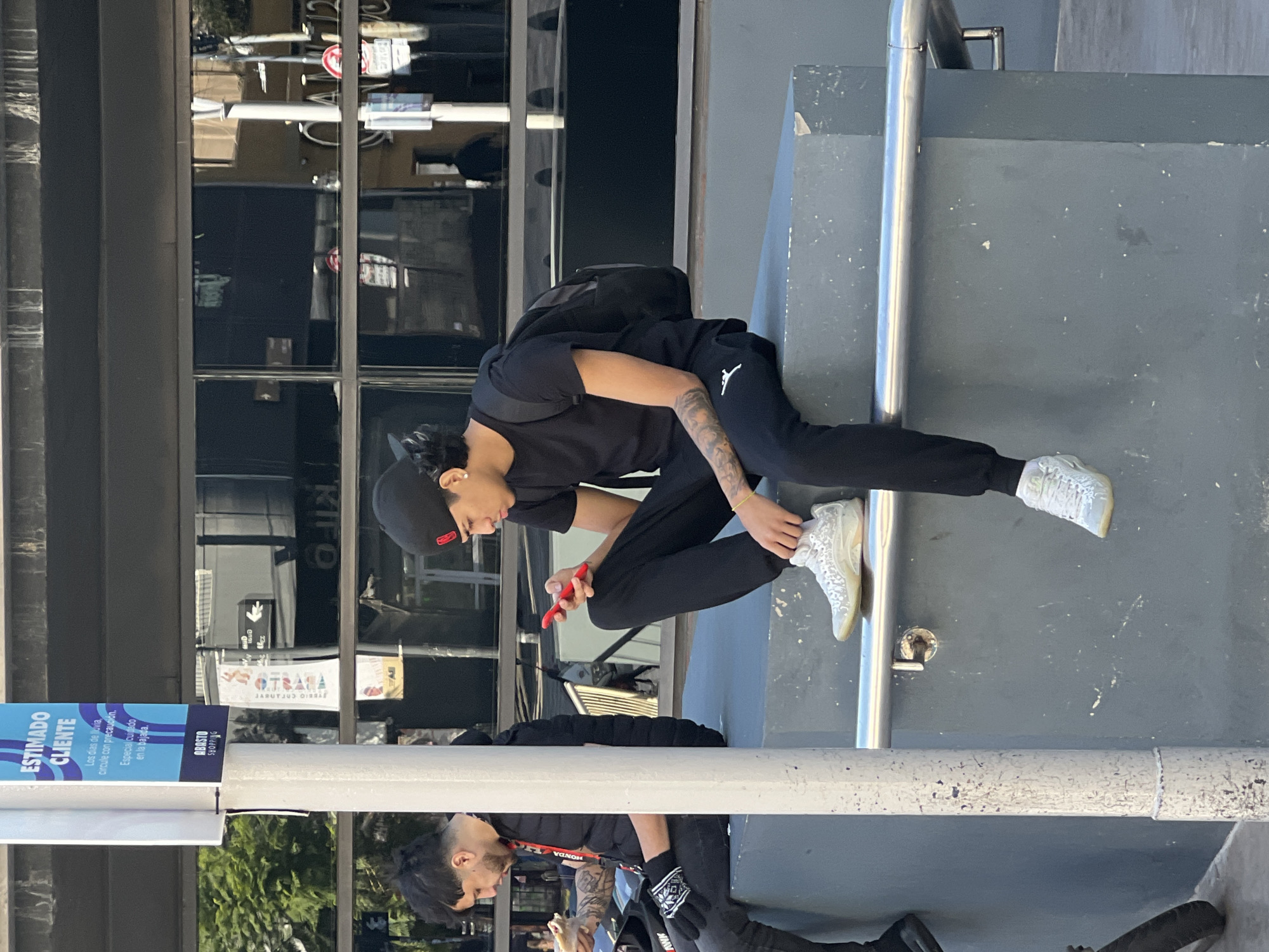 A young person wearing black clothing is sitting on a gray concrete structure next to a metal railing while holding a red cell phone. The person is wearing a black baseball cap and light-colored sneakers. Their arm has visible tattoos. Behind the person is a building with reflective windows. A sign on a pole reads "ESTIMADO CLIENTE" and mentions "ABASTO SHOPPING," indicating a location in Buenos Aires, Argentina. A reflection of a person holding what appears to be a sandwich is visible in the bottom of the image.
