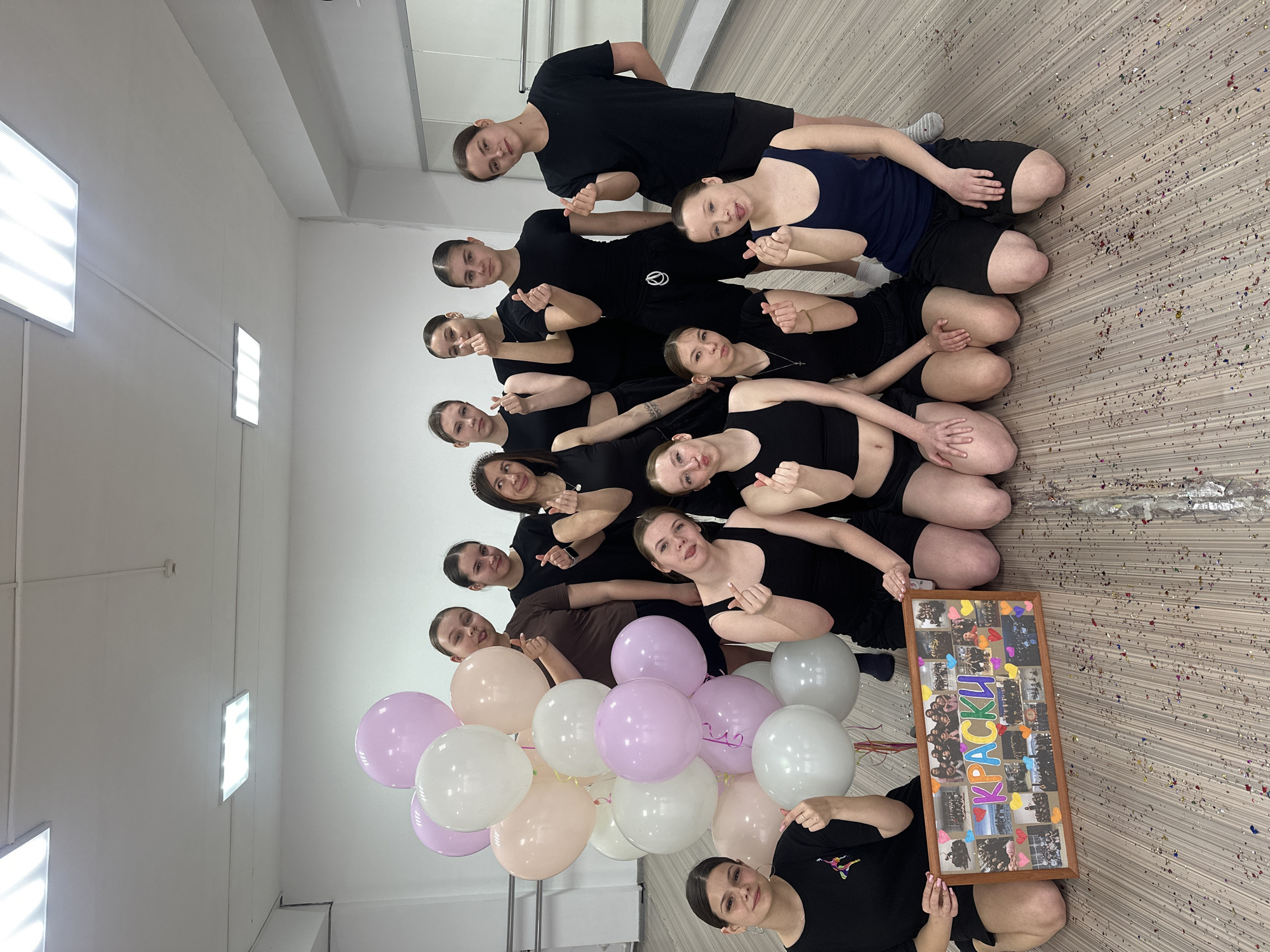 A group of 13 young women are kneeling in a row on a tiled floor in a brightly lit room. They are dressed in various black or dark-colored dance attire. Many of them are making heart shapes with their fingers. A framed collage of photos is held by one of the women, containing the word "КРАСКИ" in Russian (which translates to "Paints" in English), as well as various pictures of the group. A cluster of pastel-colored balloons lies nearby. Confetti is scattered across the floor. The scene appears to be a celebration. The city is Votkinsk, Russia, based on the wording on the collage.