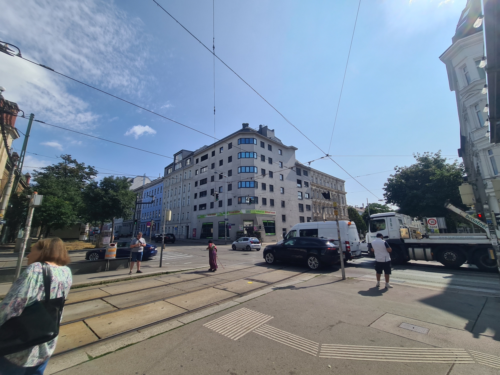 In Vienna, Austria, a sunny urban street scene unfolds at an intersection with tram tracks. A woman in a patterned blouse and black shoulder bag walks away from the camera. Across the tracks, a person in a red dress with a broom crosses the street. To the right, a black SUV is parked on the street, and a white van is visible behind it. A large white truck with "Brantner" branding and a "30 Zone" sign is parked further down the road. In the background, a multi-story building with shops on the ground floor stands prominently, and several other buildings line the street under a bright blue sky with scattered clouds.