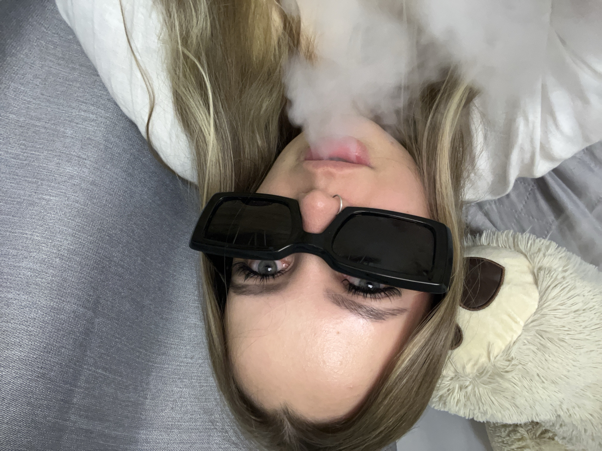 A woman with blonde hair lies on a gray surface. She is wearing black sunglasses and a nose ring, and smoke is emerging from her mouth. A white pillow and a stuffed animal are also visible in the frame. It is not possible to determine the location of the scene as being in Krasnodar, Russia.