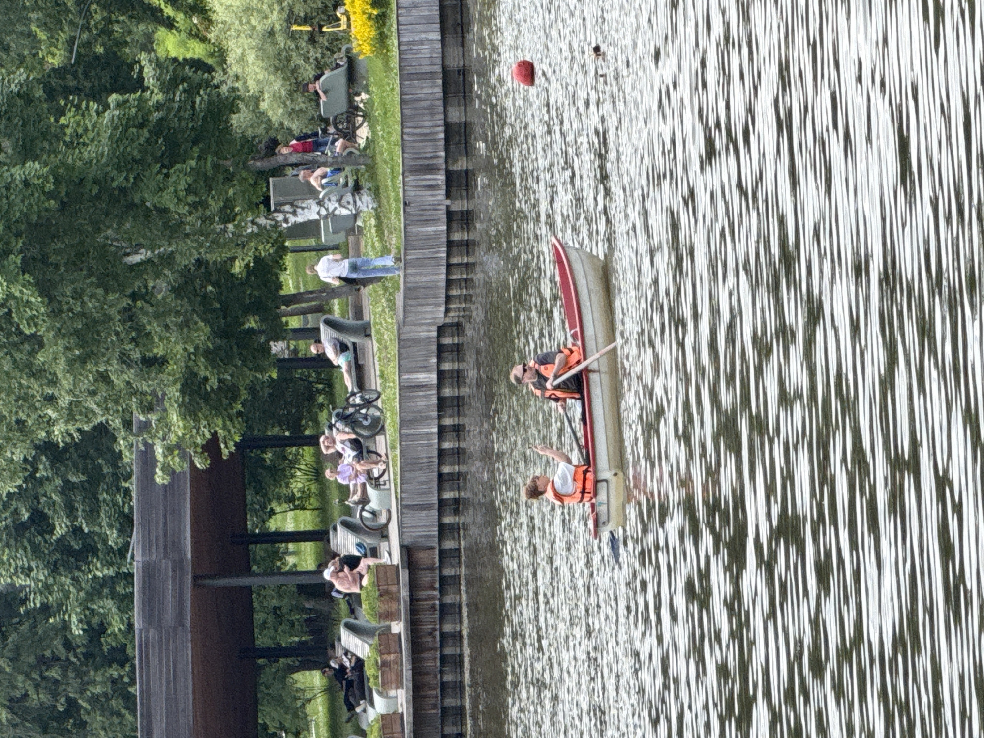 Two individuals are in a red and white rowboat on a body of water, likely a river or lake. The person in the front is steering and rowing, while the person behind them is holding up a hand, possibly pointing or signaling. Both individuals are wearing orange life vests. In the background, a wooden pier or boardwalk runs along the edge of the water, with several people relaxing on lounge chairs and benches. Some individuals are dressed in summer clothing. Behind the pier, a covered structure with a dark roof, possibly a pavilion, is visible, along with numerous trees and greenery. The scene appears to be a park or recreational area in Moscow, Russia, on a bright day.
