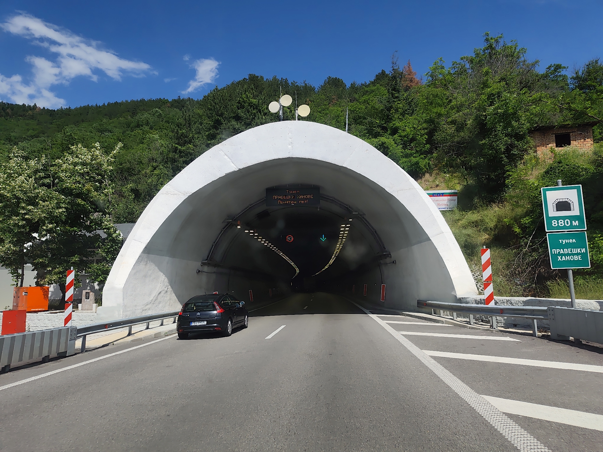 The image shows the entrance to a tunnel on a sunny day. The tunnel has a large, white, arched entrance with a road leading into it. A dark-colored car is entering the tunnel from the left lane. Above the tunnel entrance, there is a digital sign displaying text in Cyrillic. There are also traffic signs on the right side of the road, one indicating a tunnel and its length (880 m) and another with text in Cyrillic. The surrounding environment is characterized by green, tree-covered hills and a bright blue sky with some clouds. The road is asphalt with white lane markings. Safety barriers are visible along the sides of the road.
