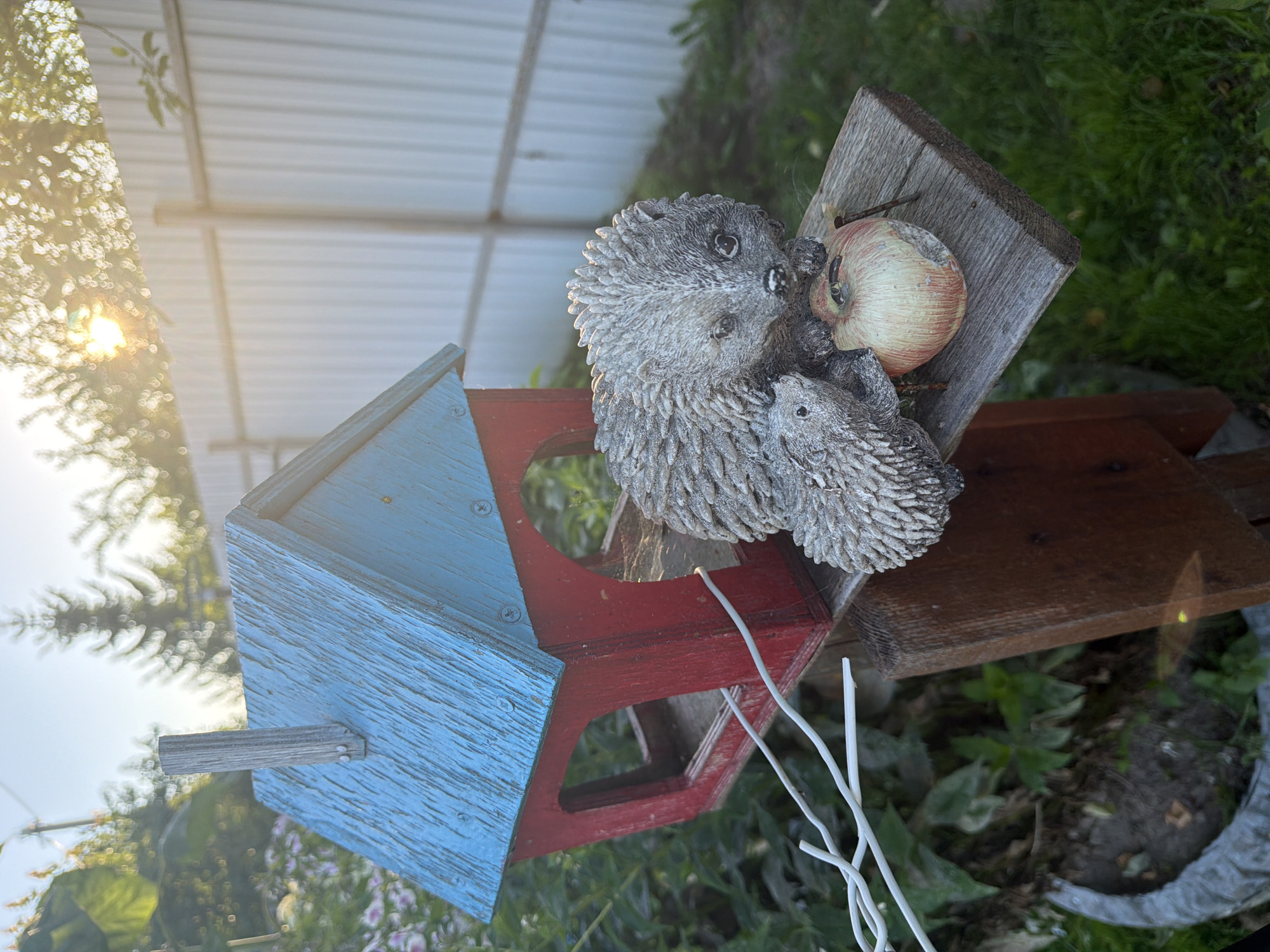 A close-up, eye-level shot captures a weathered, red and blue birdhouse in a sun-dappled outdoor setting. Two grey, textured hedgehog figurines, one larger and one smaller, rest on a wooden perch attached to the birdhouse. A single, slightly bruised apple, tinged with red and yellow, sits in front of the hedgehogs, with small twigs protruding from it. The birdhouse's blue roof shows signs of wear, and a small wooden chimney protrudes from its peak. A thin white wire snakes across the front of the birdhouse. The background features a white metal fence, lush green grass, and blurred foliage with sunlight filtering through. The scene is devoid of human presence. The image appears to be taken in the city of Primernoe, Russia, suggesting a residential or garden environment.