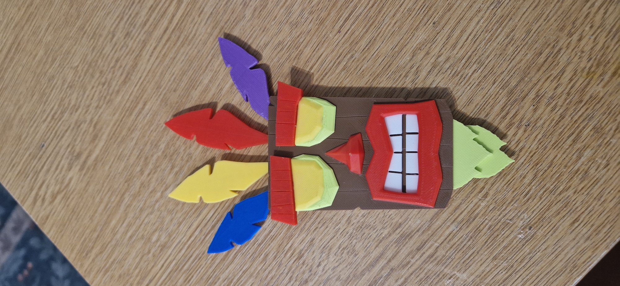 This media file depicts a brightly colored, 3D-printed object resembling a stylized tiki mask. The mask has a brown, wood-like base with several prominent features. On the left side, there are five layered, feather-like protrusions in vibrant colors: purple, red, yellow, yellow, and blue, from top to bottom. The mask itself features a central red, jagged frame with a white, grid-like interior that appears to represent a mouth with teeth. Above the mouth-like feature, there are two pairs of light green and yellow rectangular shapes, possibly representing eyes or decorative elements. To the right of the central structure, a light green, torn-edged shape protrudes, possibly representing a leaf or additional decoration. The object is positioned on a wooden surface with a distinct grain pattern. The lighting suggests an indoor setting, possibly under artificial light, as there are no strong shadows indicative of natural sunlight. No people or text are visible in the image. The object's design and vibrant colors suggest it might be a decorative item, a toy, or a component of a larger display. The setting appears to be a table or desk in a domestic or workshop environment, given the wooden surface and the nature of the object. The overall impression is one of a playful and creative handmade or 3D-printed item.