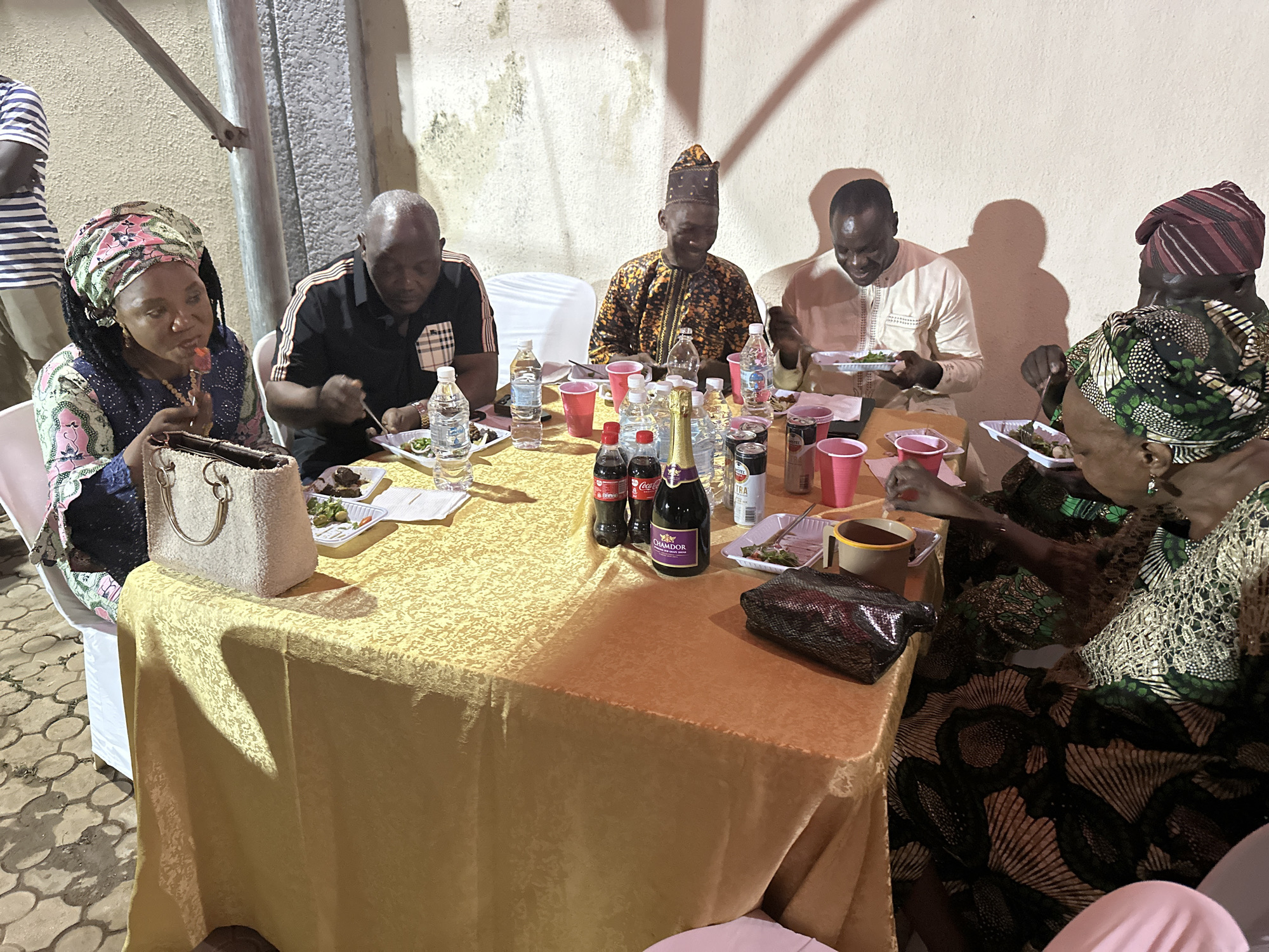 A group of six people are seated around a rectangular table covered with a gold-colored tablecloth at an outdoor event, potentially a celebration, in what may be the city of Narayi, Nigeria. They are eating from plates of food. The people are dressed in a mix of modern and traditional African clothing and headwear. The table is cluttered with food, plastic cups, bottles of water, Coca-Cola, and a bottle of sparkling wine. There are bags placed on the table as well. A pole is seen on the top left and a white wall in the background.